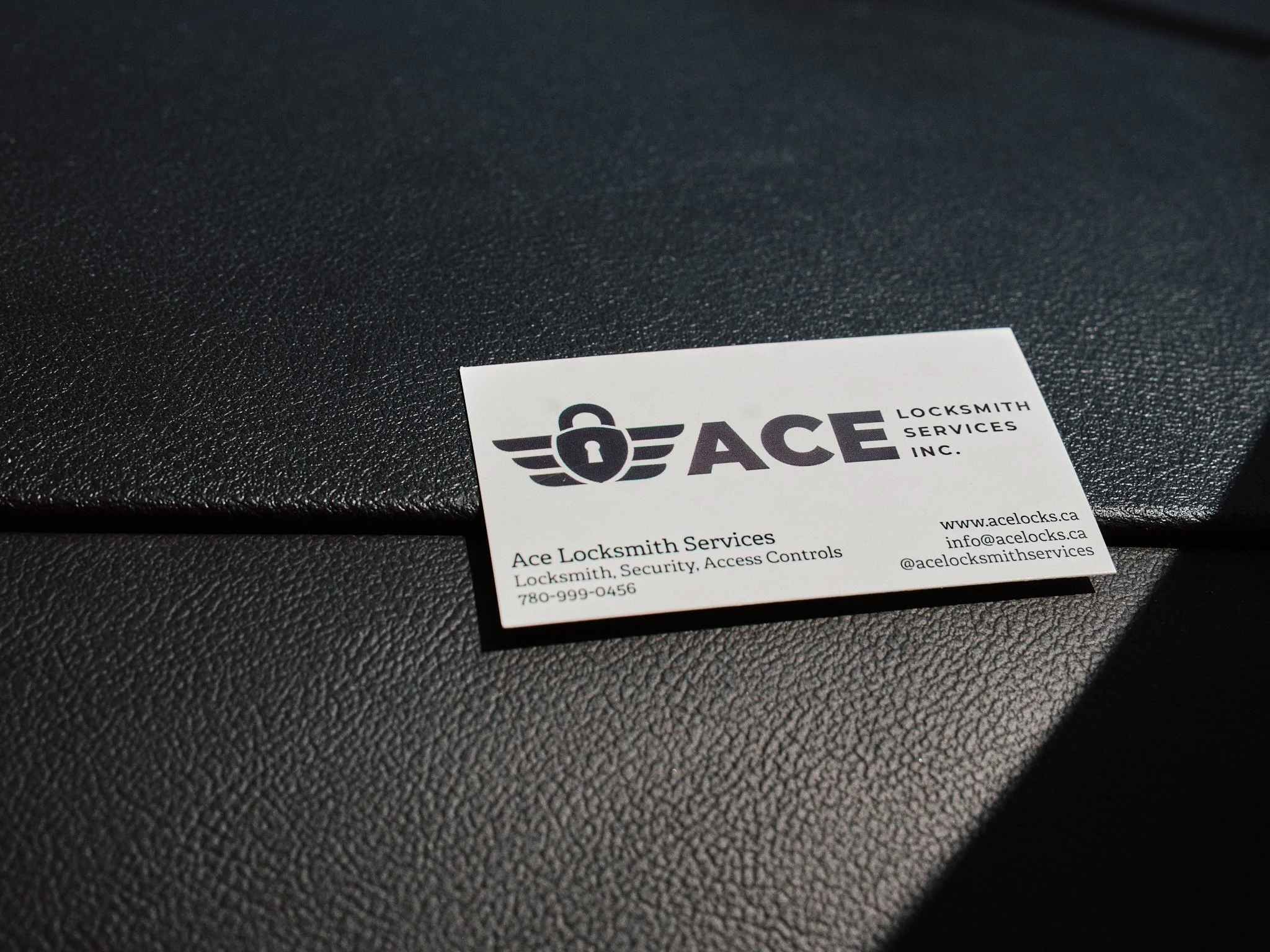 Close-up of a business card for Ace Locksmith Services Inc. on a textured black surface. The card features a logo with a lock and wings, and includes contact information such as website, email, and phone number.
