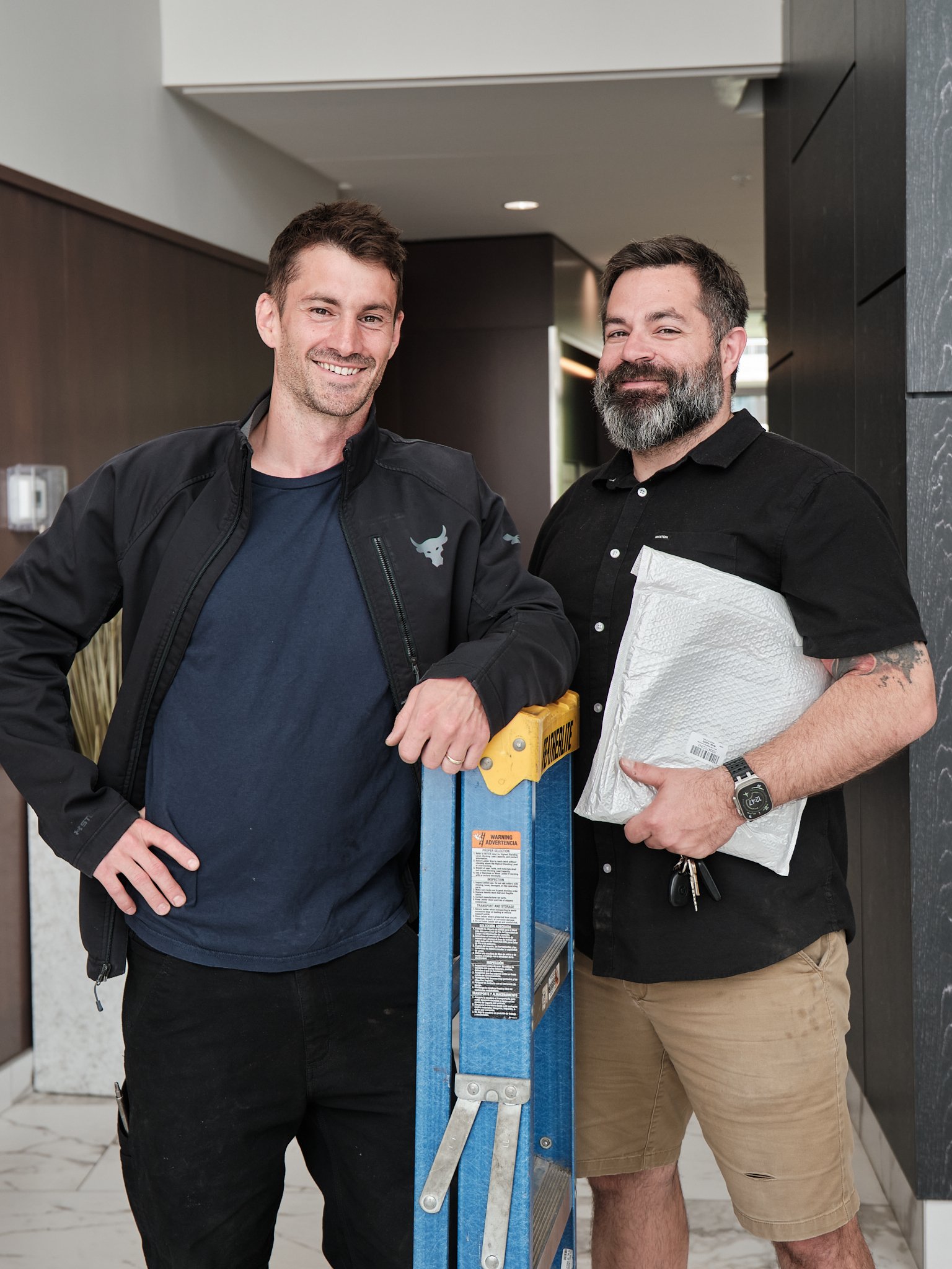 A locksmith standing next to one of his property manager clients. Both men are smiling with the locksmith holding a blue ladder and the property manager holding documents.