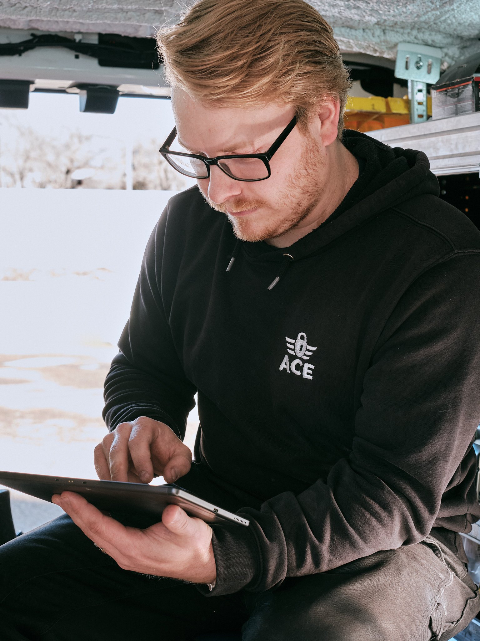 A man with light brown hair, glasses, and a beard is sitting outdoors, holding and looking at a tablet device. He is wearing a black hoodie with a logo on the chest and beige pants. The background shows snow on the ground and some trees in the distance.
