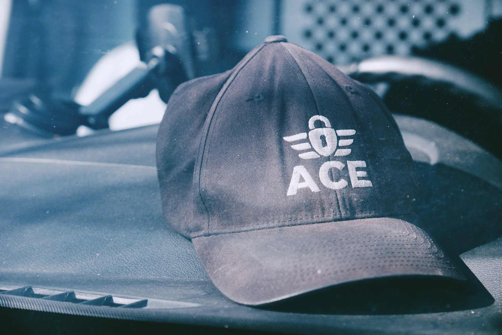 A black cap with a white logo reading 'ACE' and a circular emblem on the front, resting on a car dashboard. Ace Locksmith Services Inc, located in Edmonton, Alberta.