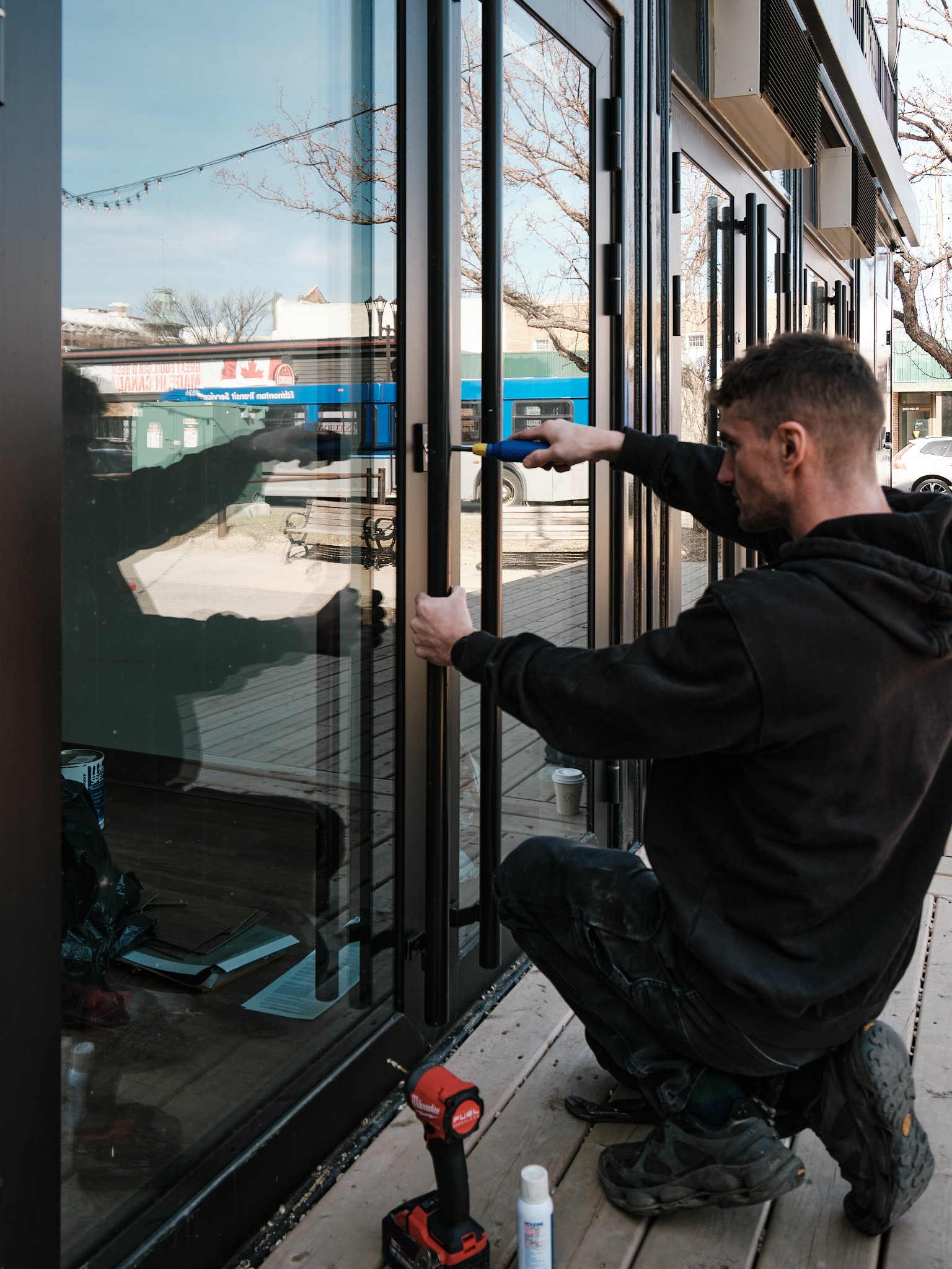 The owner of Ace Locksmith Services Inc. kneeling on a wooden deck installing or repairing a glass door using a screwdriver, with tools and materials around him.