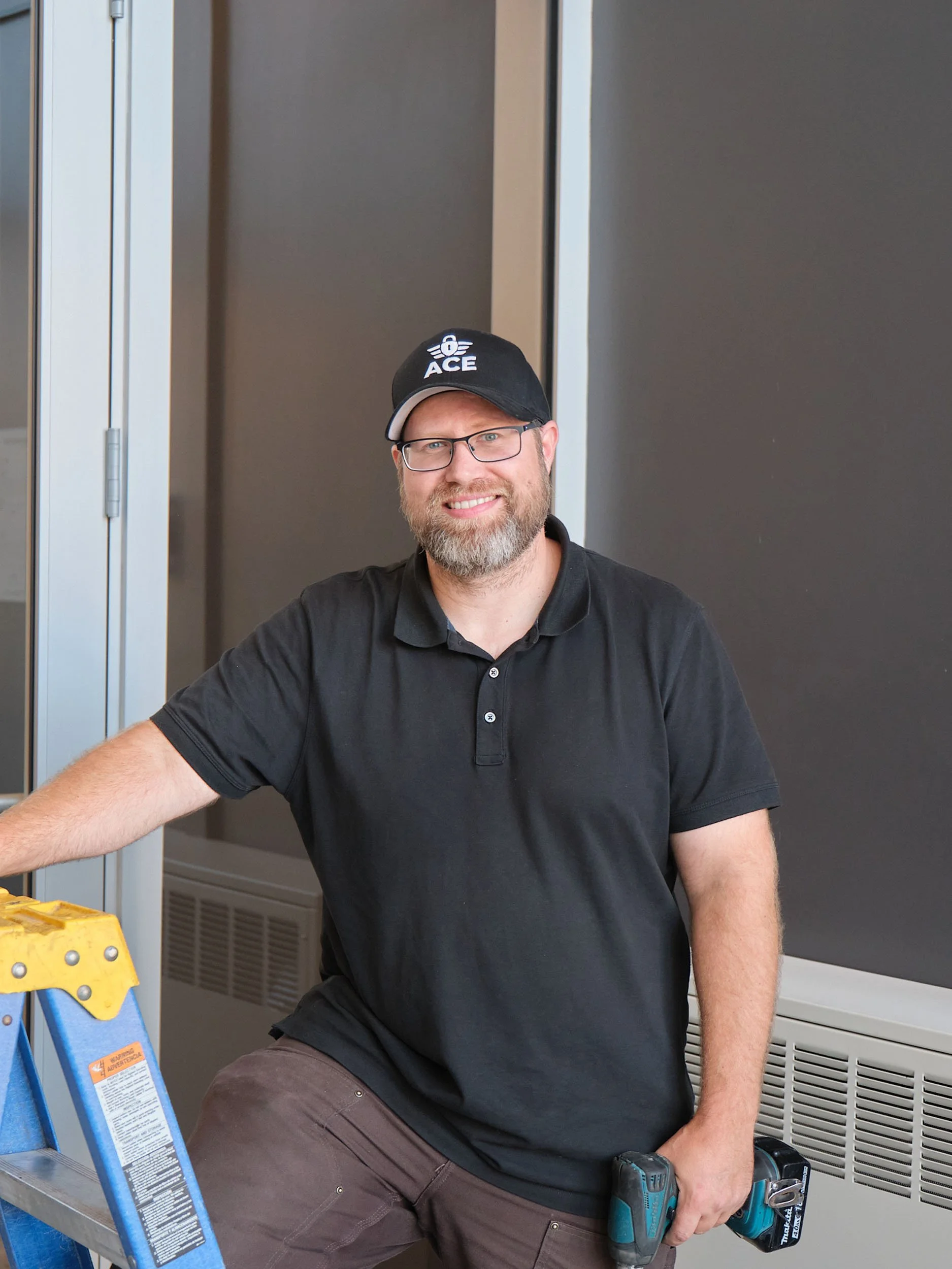 A man wearing glasses, a black polo shirt, a black cap with a logo, and brown pants is kneeling on one knee, holding a cordless electric drill, next to a yellow step ladder, in an indoor setting with a dark wall and a metallic door frame.
