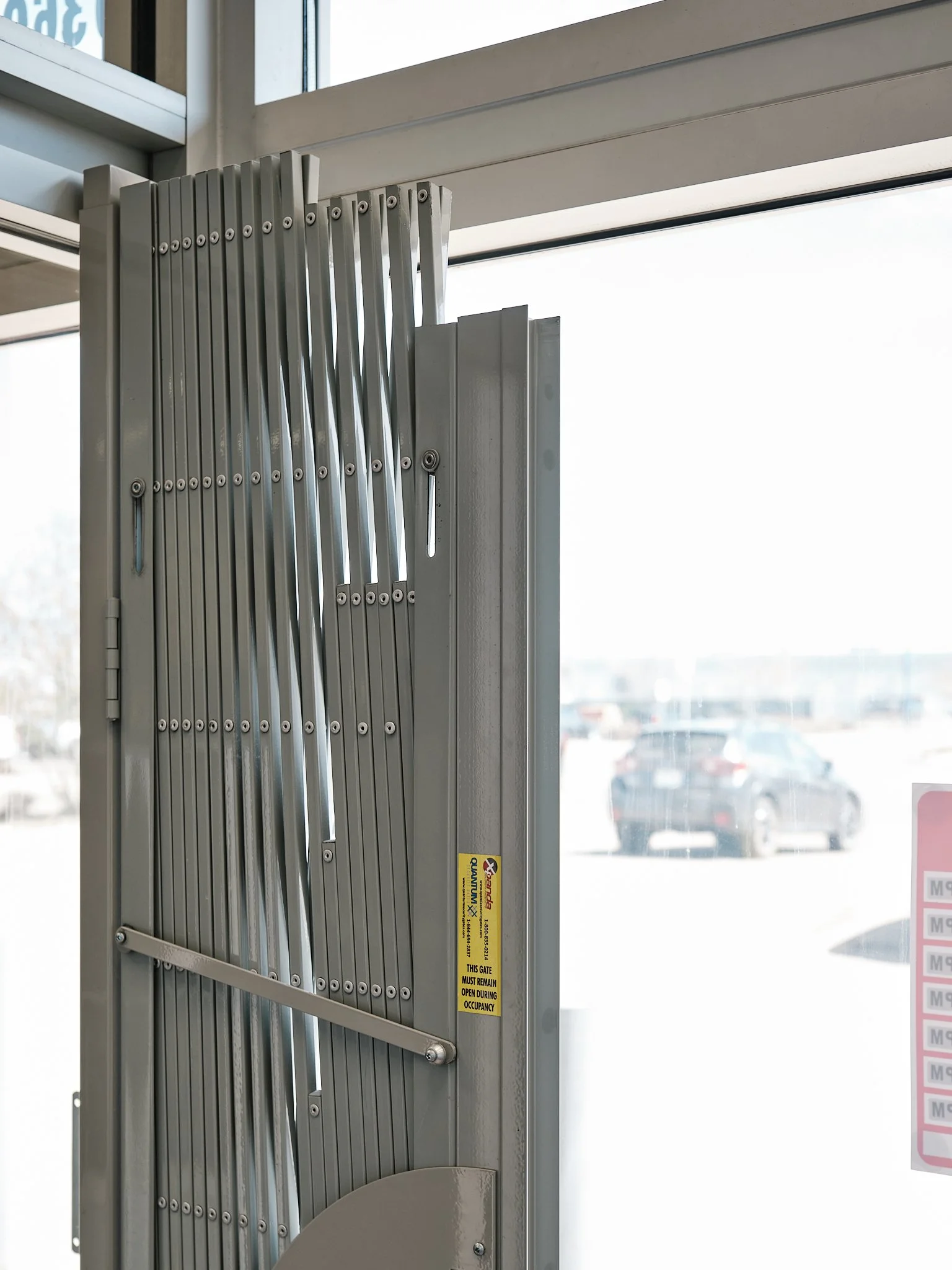 Security gate with metal bars in front of large glass window, with parking lot visible outside.