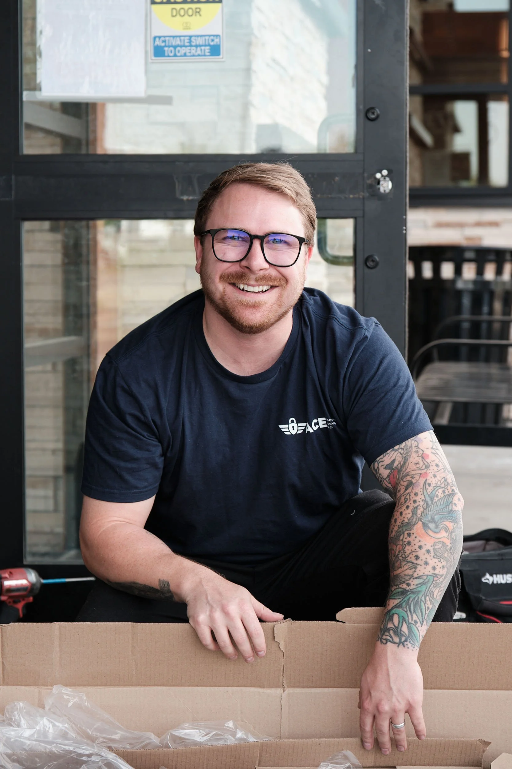 A smiling man with glasses and tattoos on his right arm, wearing a black T-shirt, is sitting at a table with a cardboard box in front of him. The scene appears to be in an outdoor or semi-outdoor setting with a brick wall and windows in the background, and a yellow and blue safety sign on the window.