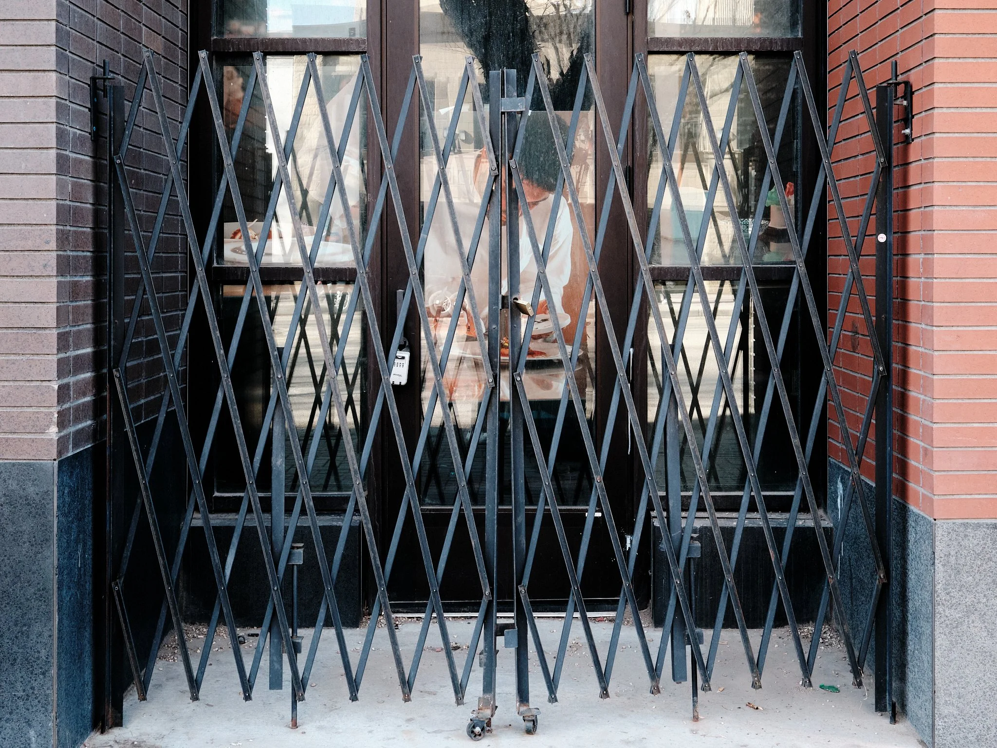 A person standing inside a building behind a closed black metal accordion-style security gate, with glass windows and brick walls on each side.
