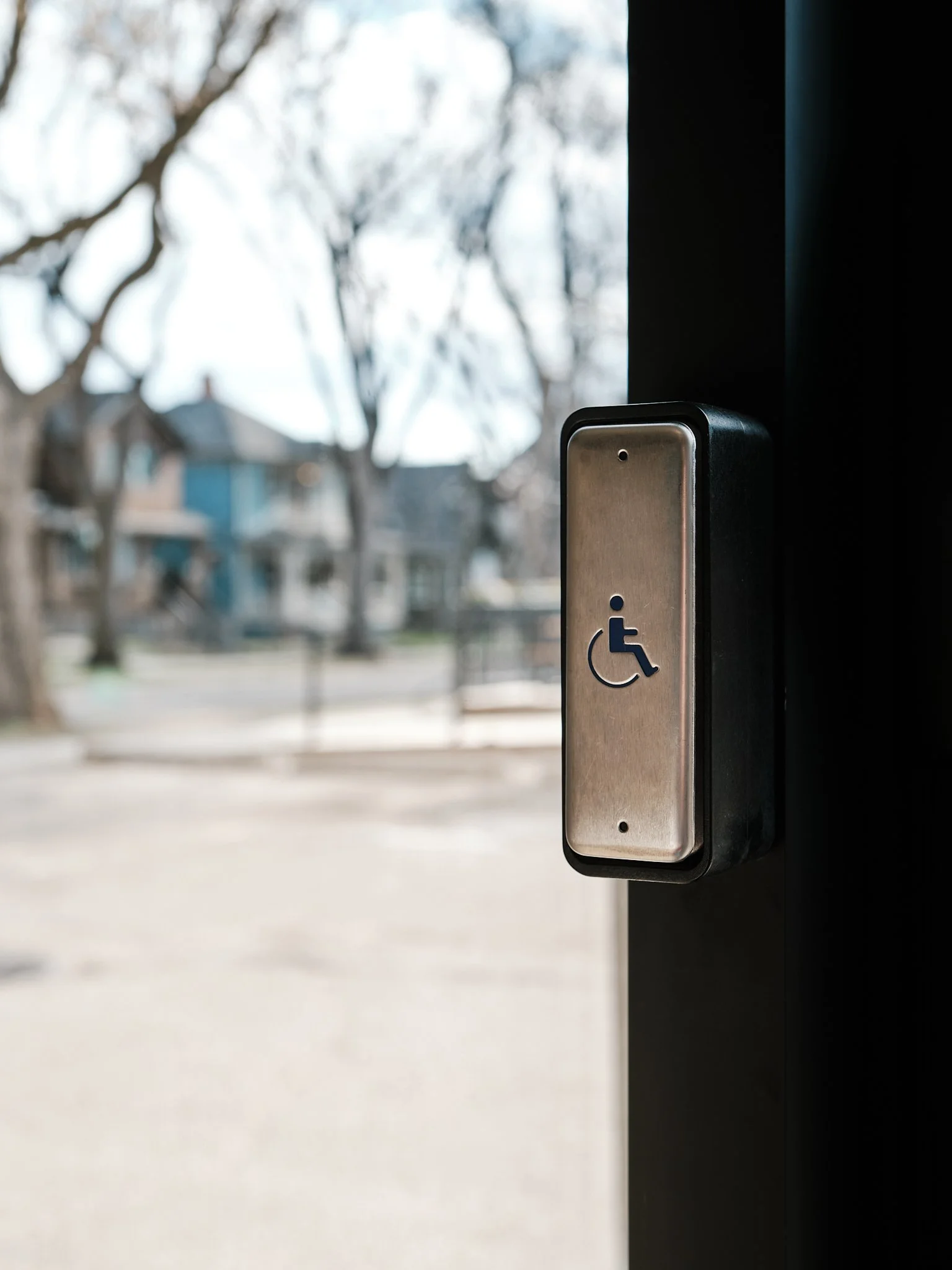 Close-up of a wheelchair accessible button on a wall near a door, with residential buildings and leafless trees in the background, viewed through a window.