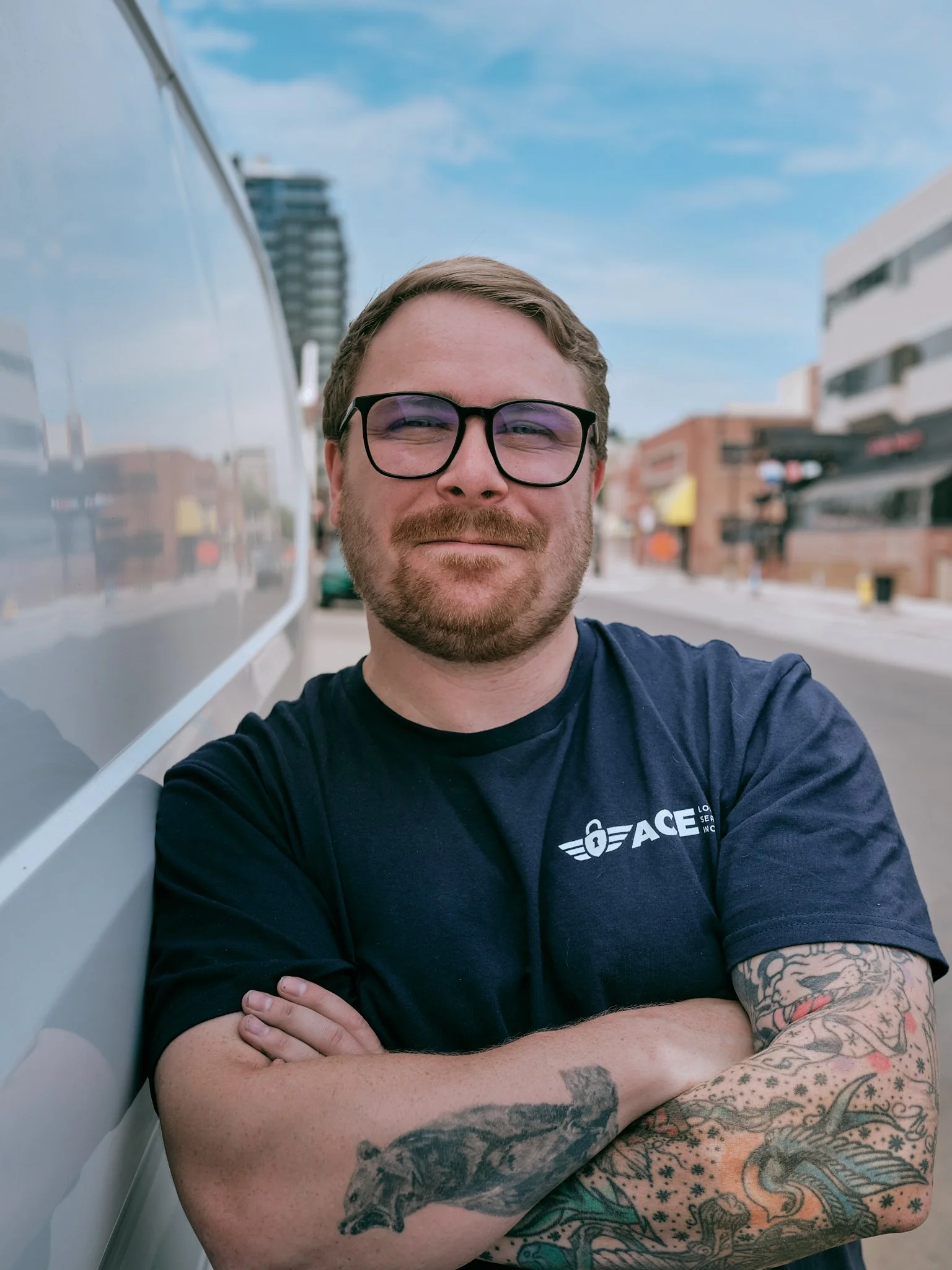 A man with glasses and tattoos on his arms standing outdoors with arms crossed, smiling at the camera, with buildings and a blue sky in the background. Ace Locks employee.