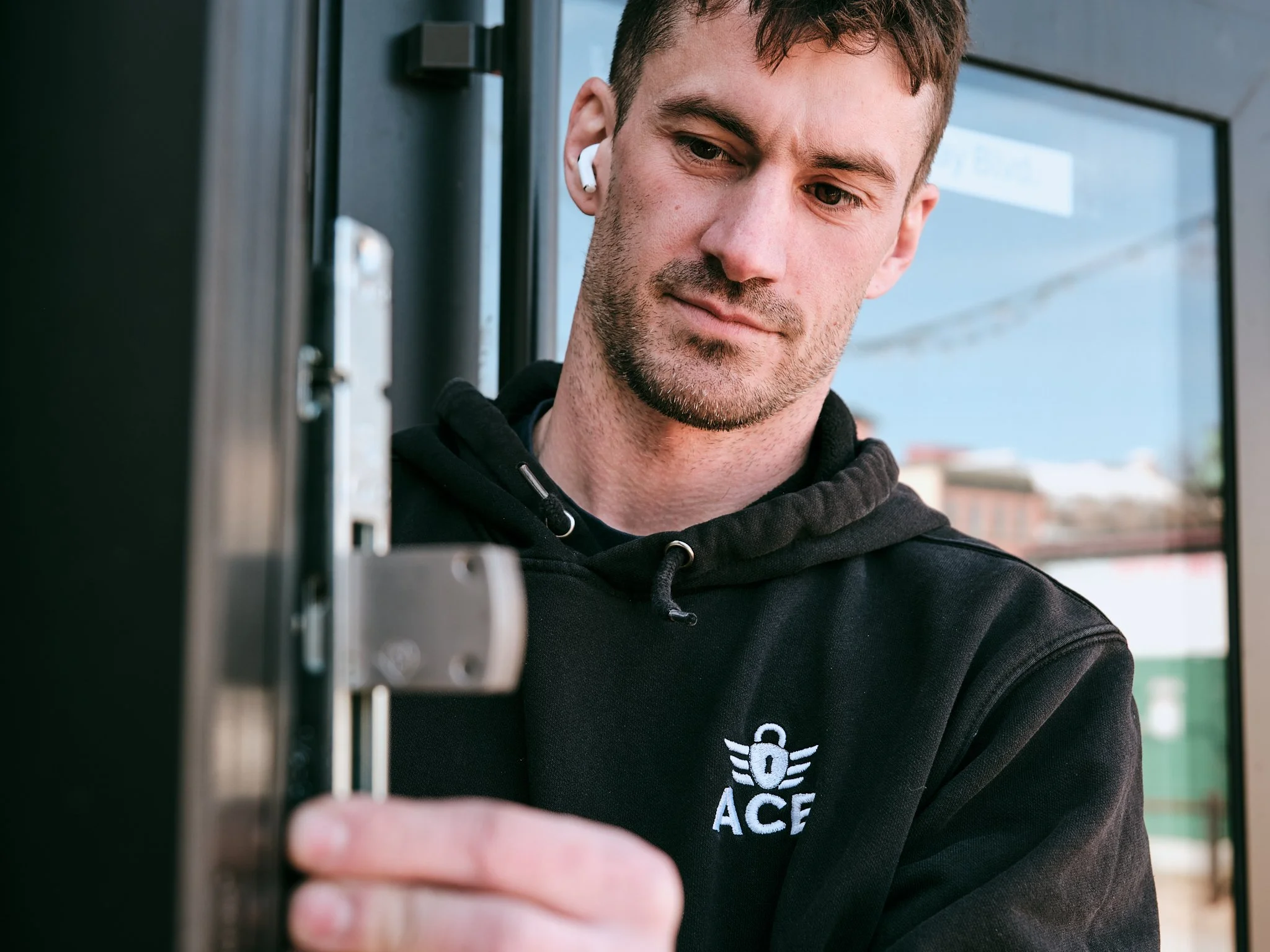 The owner of Ace Locksmith Services inc. A man with short brown hair, facial stubble, and AirPod wireless earbuds in his ears, wearing a black hoodie, is smiling slightly while holding a commercial lock he is about to install.