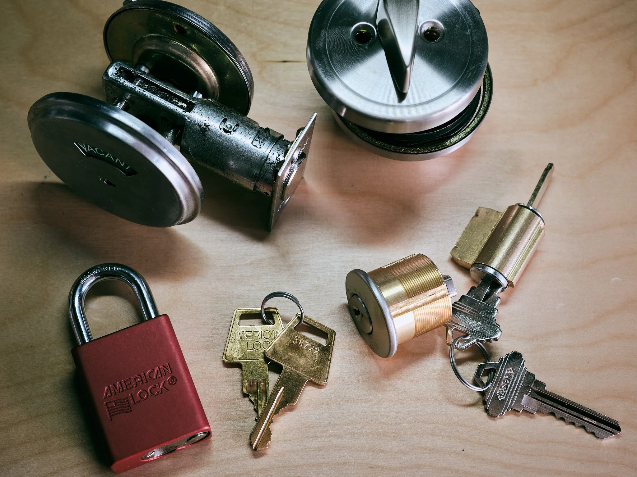 A collection of locks, keys, and security devices arranged on a wooden surface. Includes a red American Lock padlock, several keys, and various metal locks and components.