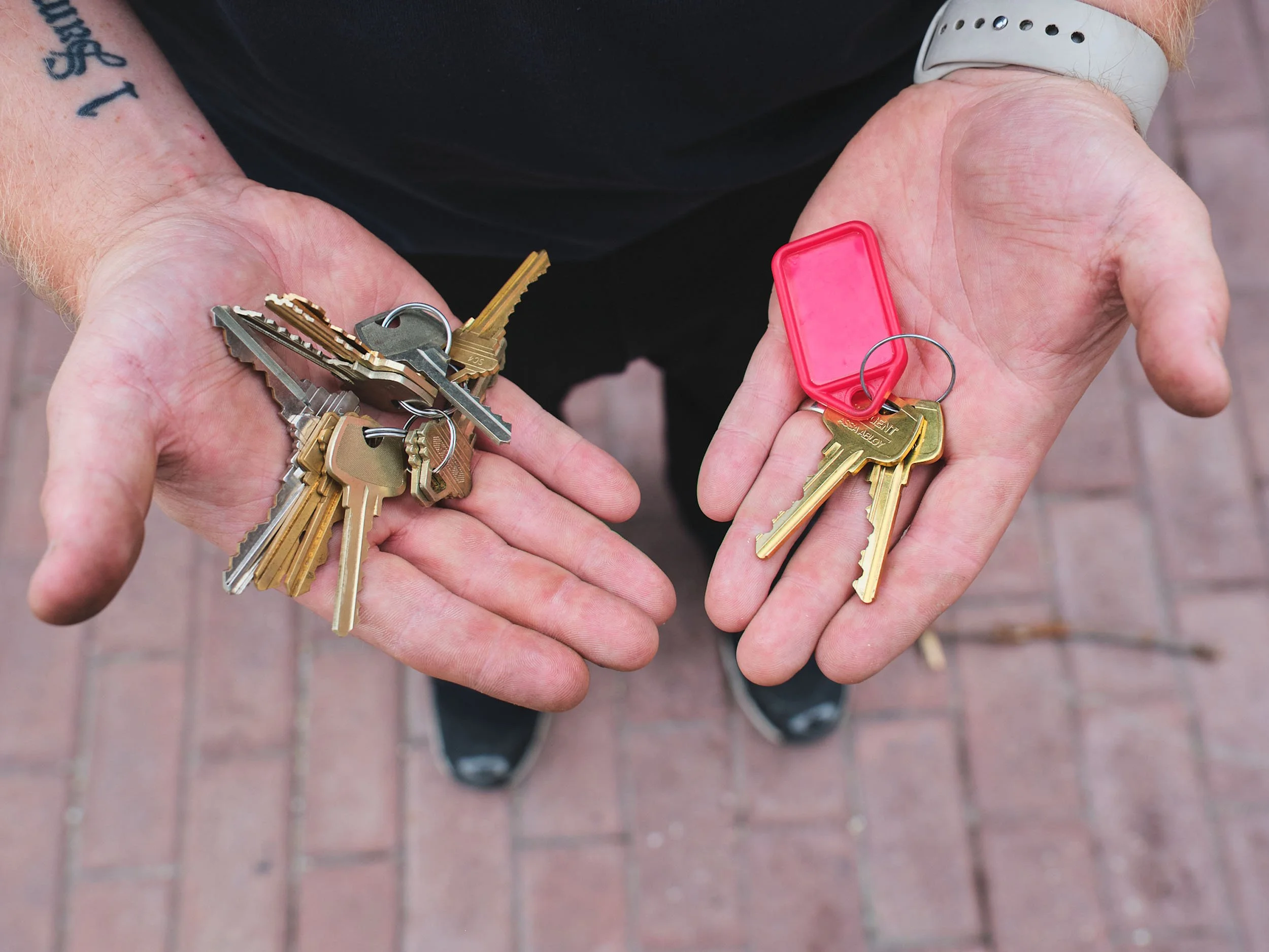 A person holding a bunch of keys with a red key fob, wearing a white bracelet and standing on a brick sidewalk.