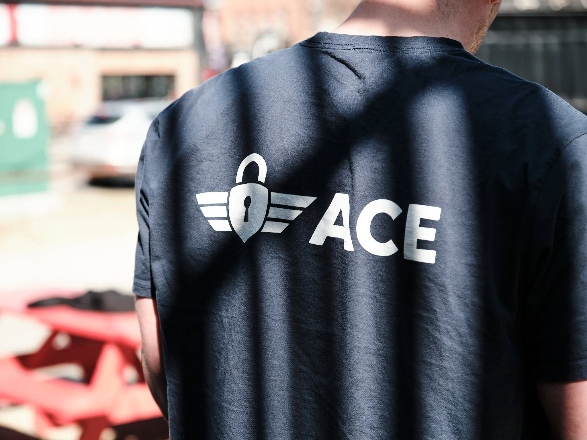 Person wearing a dark blue uniform with a logo that says 'ACE' and a wings insignia on the back, standing outdoors near a red fence. He is wearing the t-shirt for Ace Locksmith Services, a locksmith in Edmonton, Alberta.