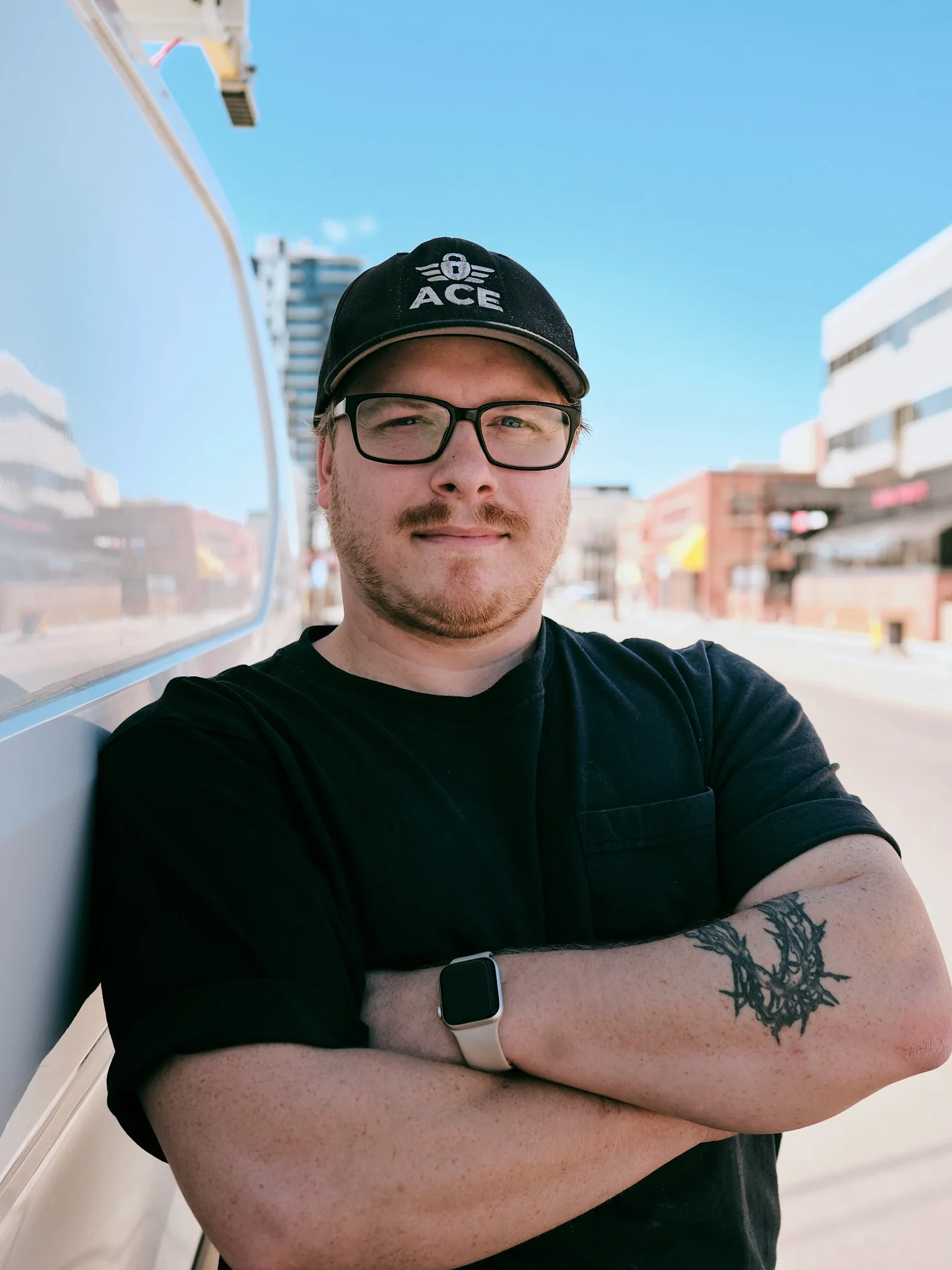 A certified locksmith with Ace Locksmith Services Inc. A young man with glasses and a black cap with the word 'ACE' and a winged emblem, smiling with arms crossed, standing outdoors on a sunny day with city buildings in the background.