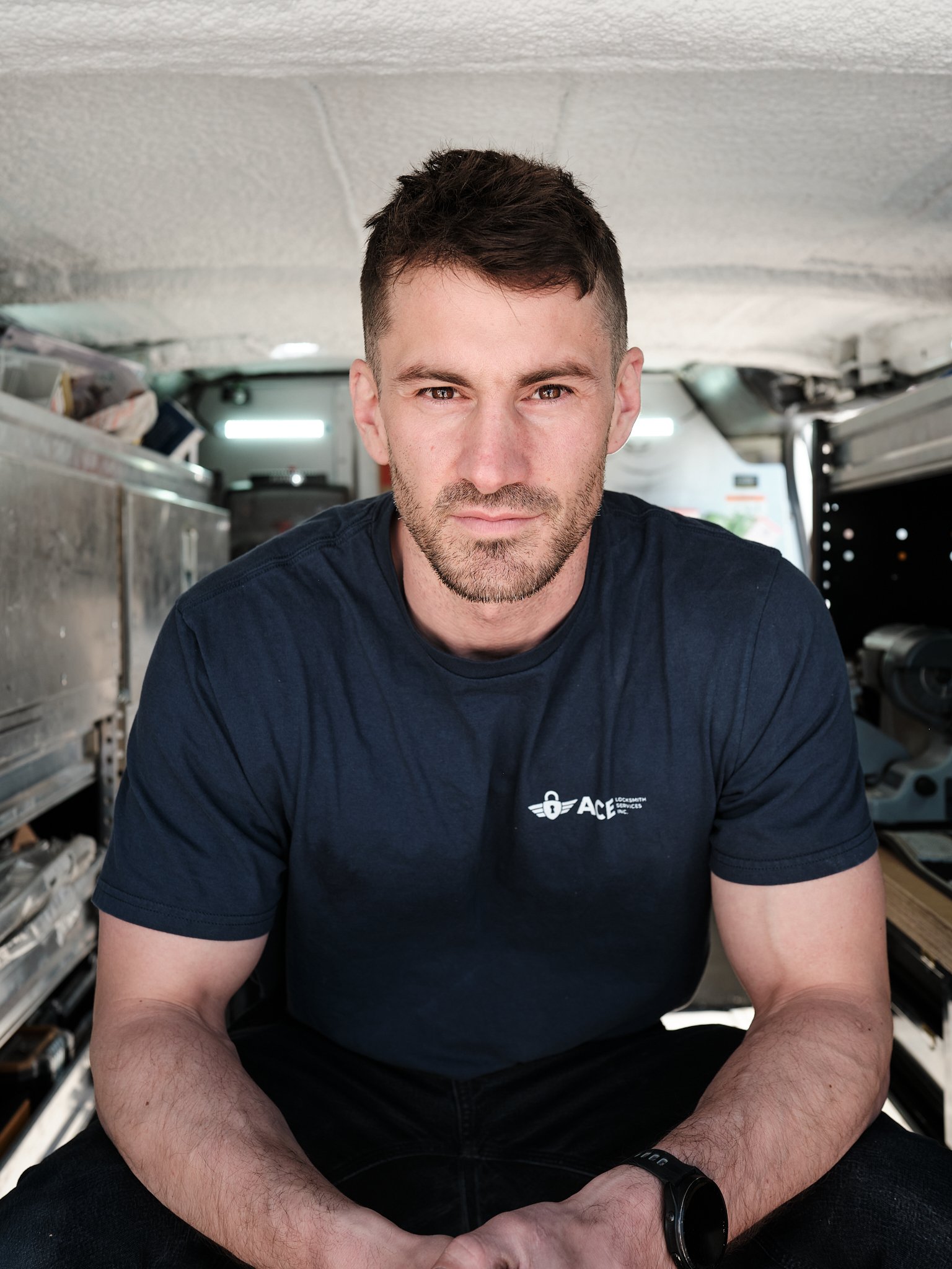 A man with short dark hair and a beard looking directly at the camera, wearing a black T-shirt with a logo that says 'ACE Locksmith Services Inc.', a commercial and residential Edmonton locksmith company.