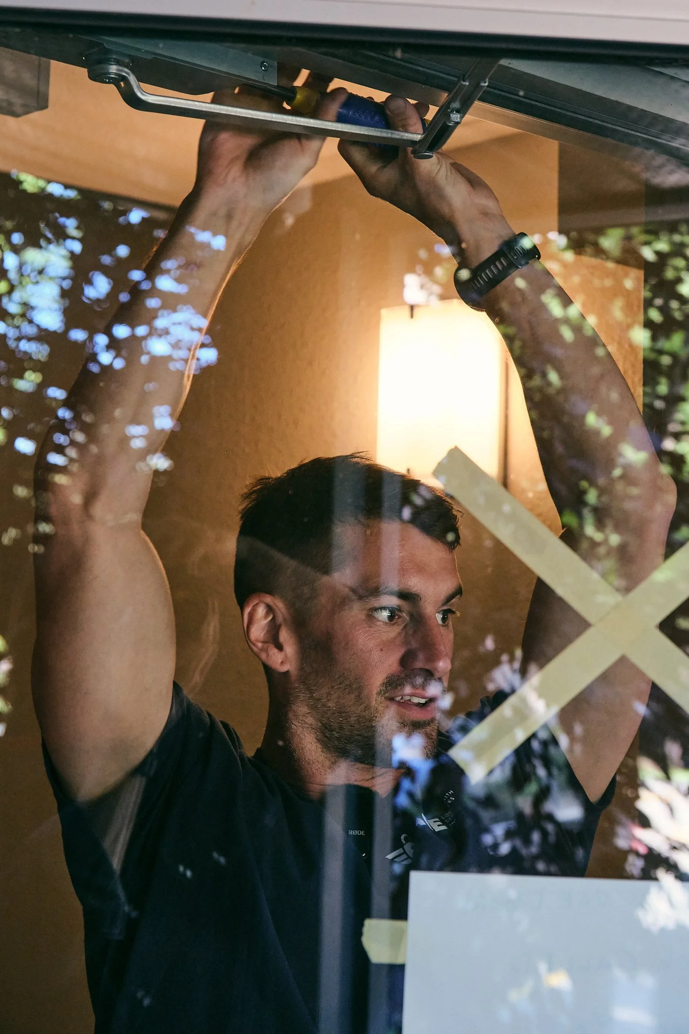 A man with short hair and a beard is behind a glass display, holding an object above his head with both hands. Reflections of trees and light are visible on the glass.