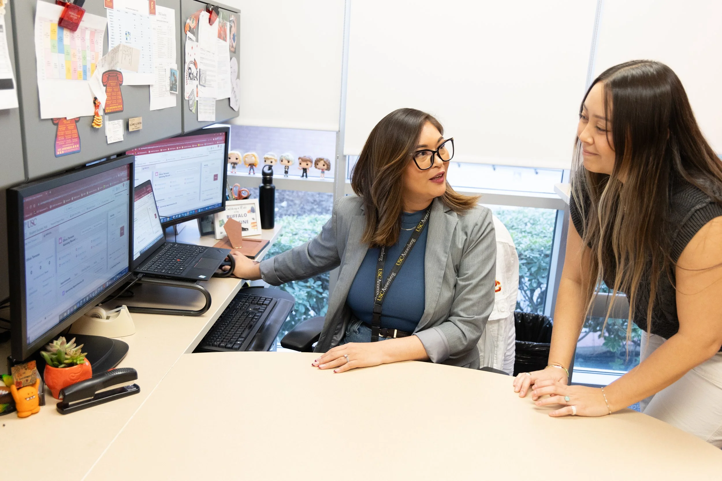 Educational counselor and student in an office, counselor seated at a desk with dual monitors and student standing and leaning on the desk, engaged in conversation.