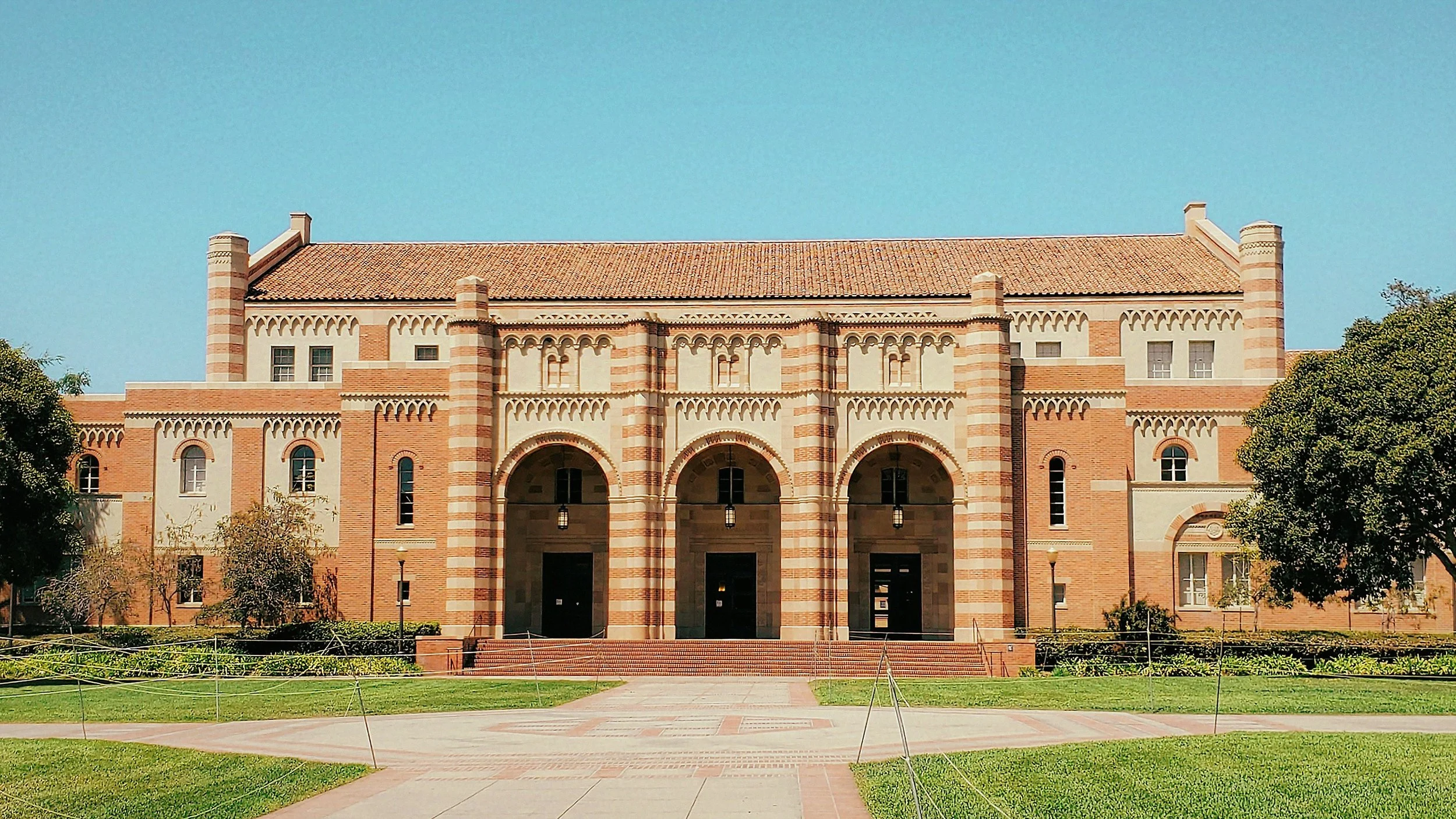A historic brick building with three large archways at the entrance, surrounded by well-maintained grass and trees, under a clear blue sky.