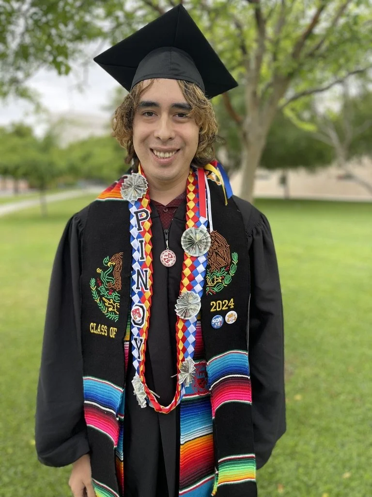 Graduating student in black cap and gown, adorned with colorful honor cords, medals, and a Mexican blanket stole, standing outdoors on green grass with trees in the background.