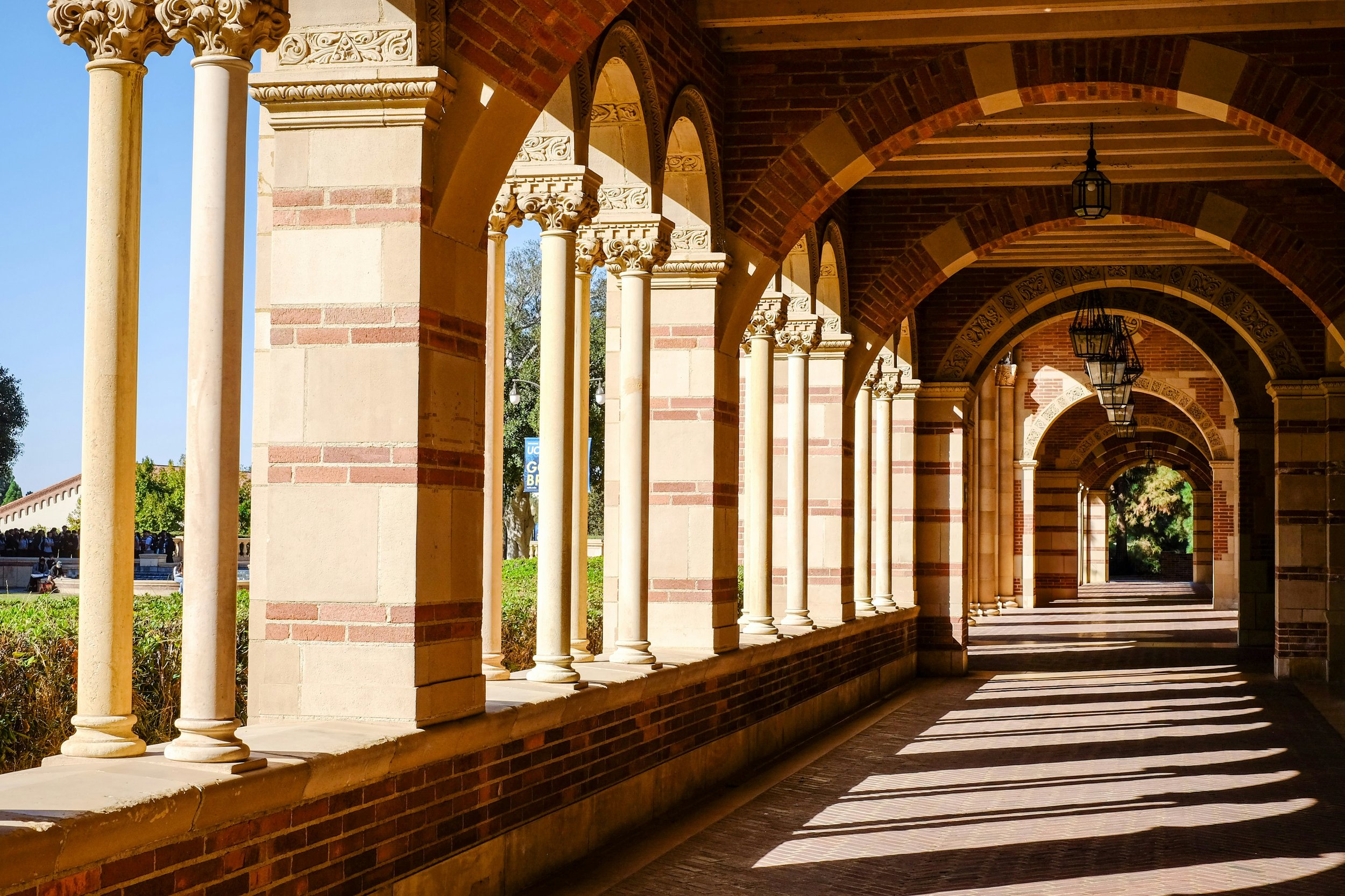 A covered walkway with brick and stone columns, arches, and hanging lanterns, with sunlight casting shadows on the floor.