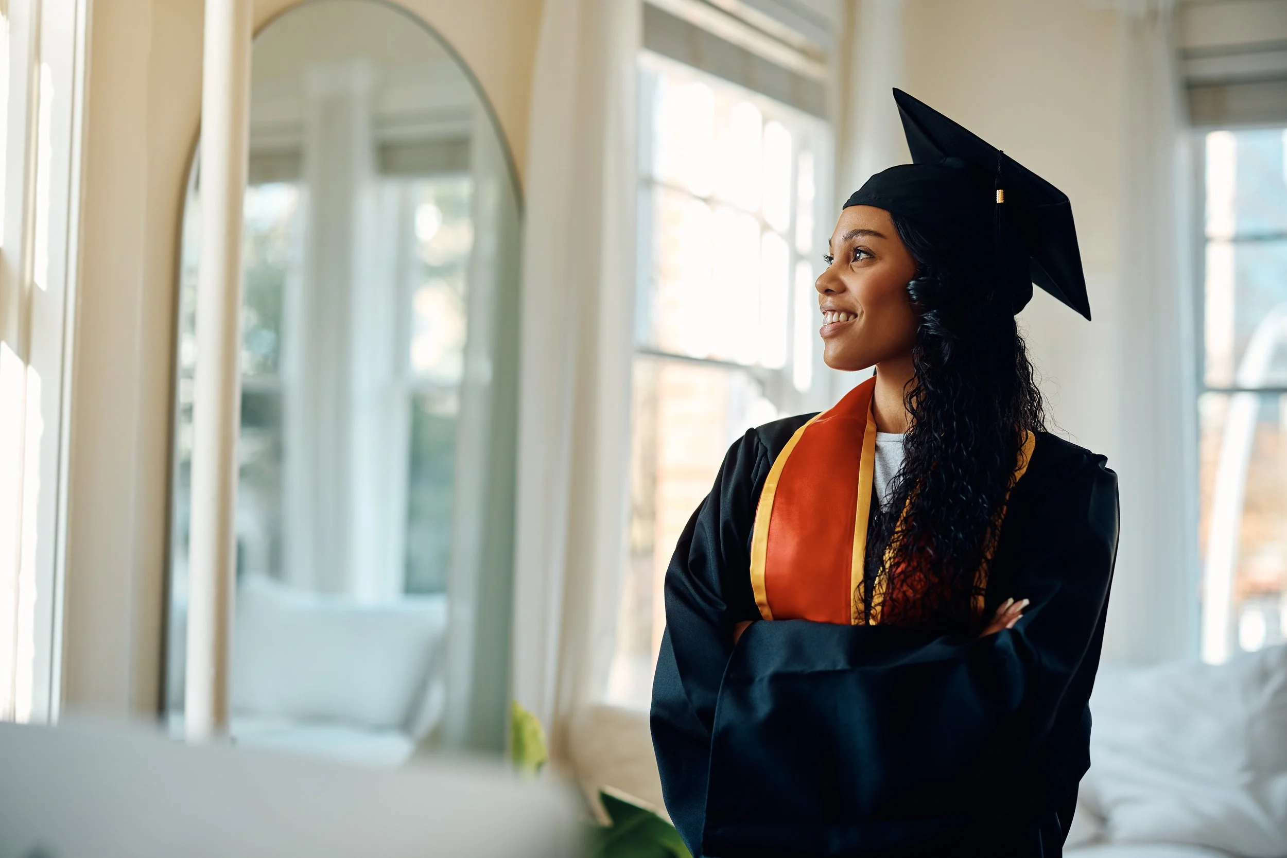 A young woman wearing a graduation cap and gown stands inside a bright room with large windows, smiling and looking to her left.
