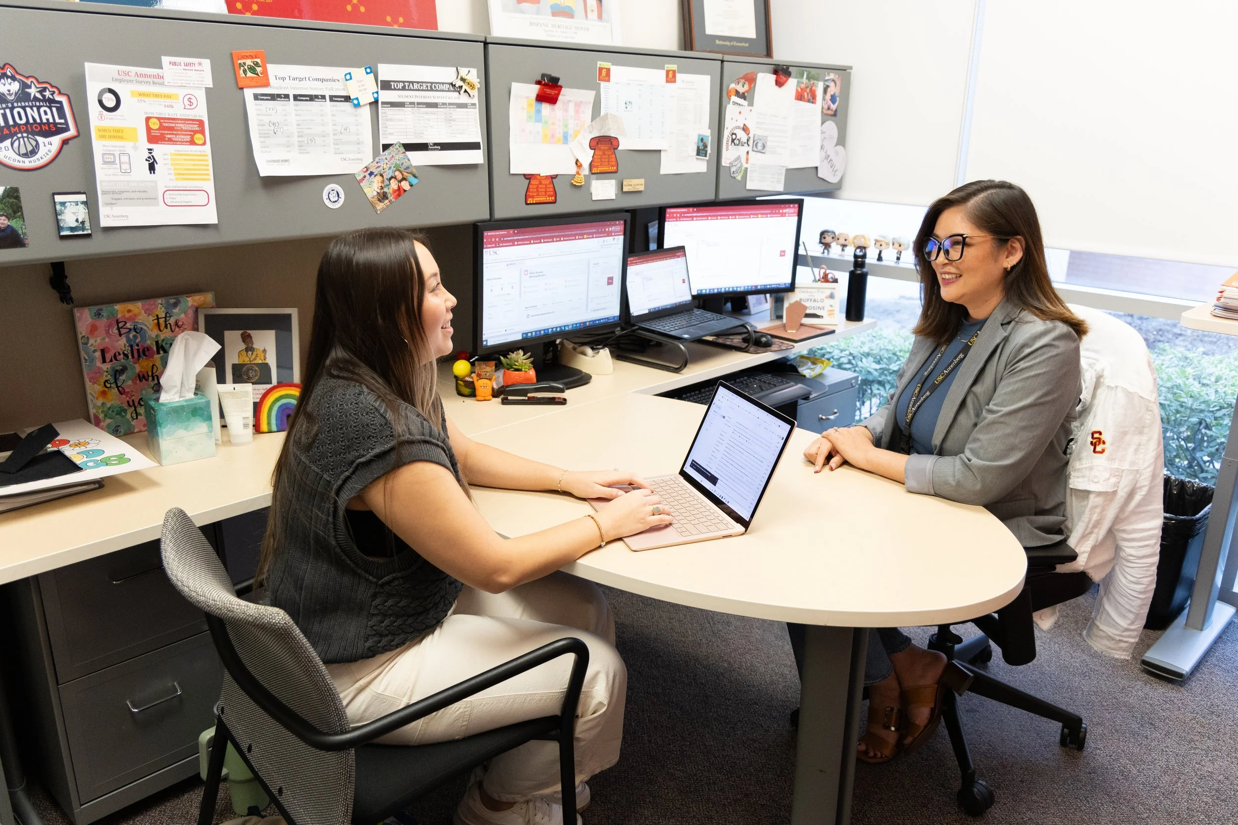 Educational counselor and student in an office, engaging in a discussion and smiling. The desk has multiple monitors, a laptop, and office supplies. The background features bulletin boards with papers and decorations.
