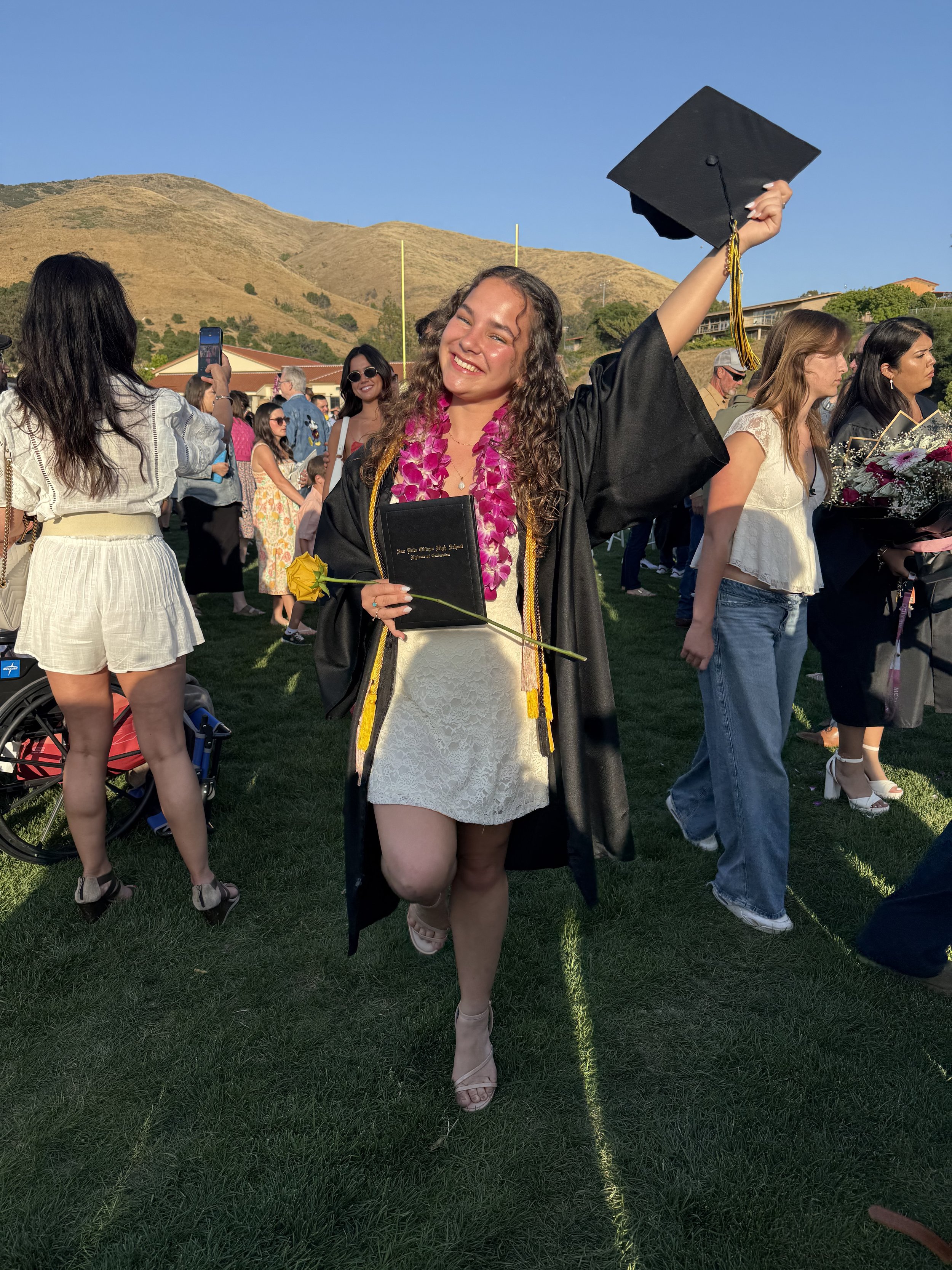 Student graduation gown and cap celebrating at outdoor graduation ceremony, holding a diploma and a yellow flower, with friends and family.