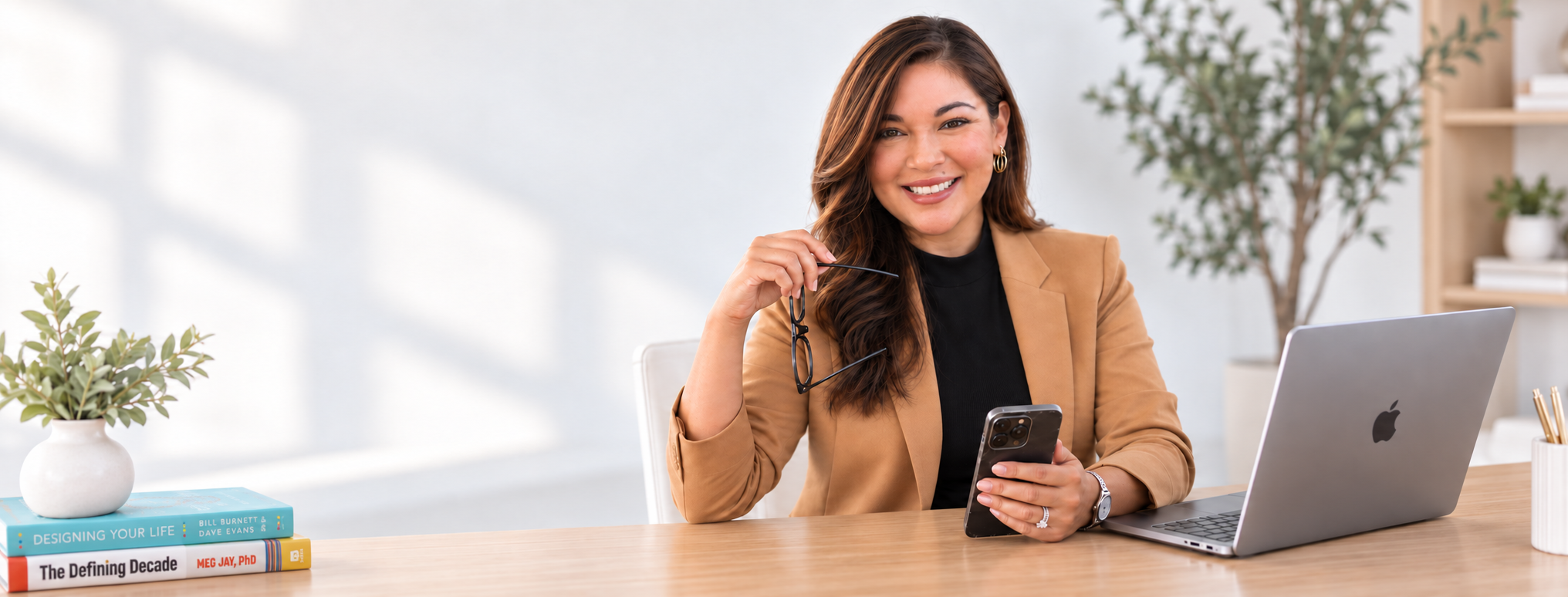 Zada Clausen, independent college admissions counselor in Los Angeles, at her desk at Zada Admissions, holding glasses in one hand and a phone in the other, smiling at the camera. There are books, a potted plant, and a modern office background.