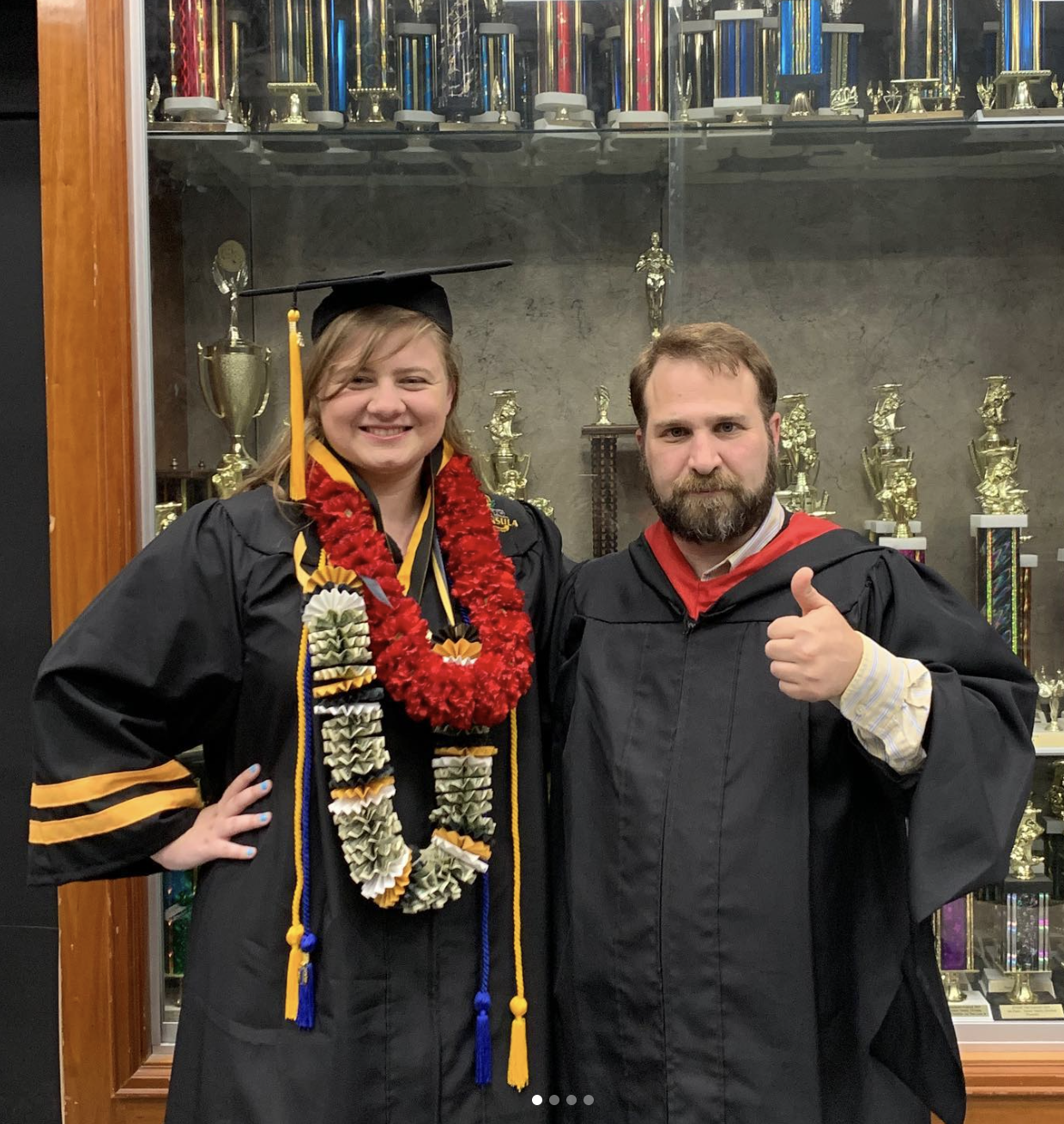 A student in a graduation cap and gown, decorated with leis, stands beside a man in a graduation gown giving a thumbs up. They are inside a trophy display case.