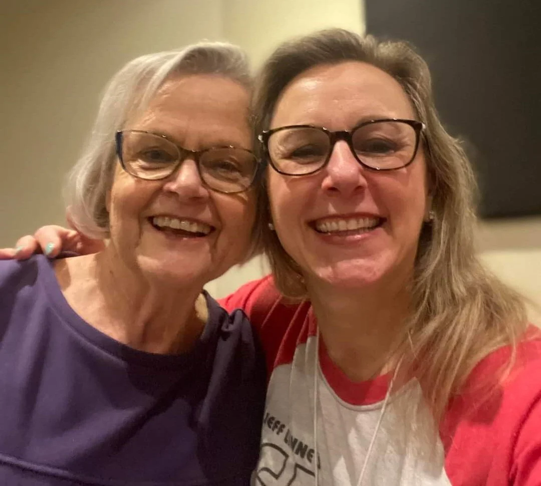 Two women smiling for a selfie, one elderly with gray hair and glasses and one middle-aged with brown hair and glasses, both wearing casual tops.