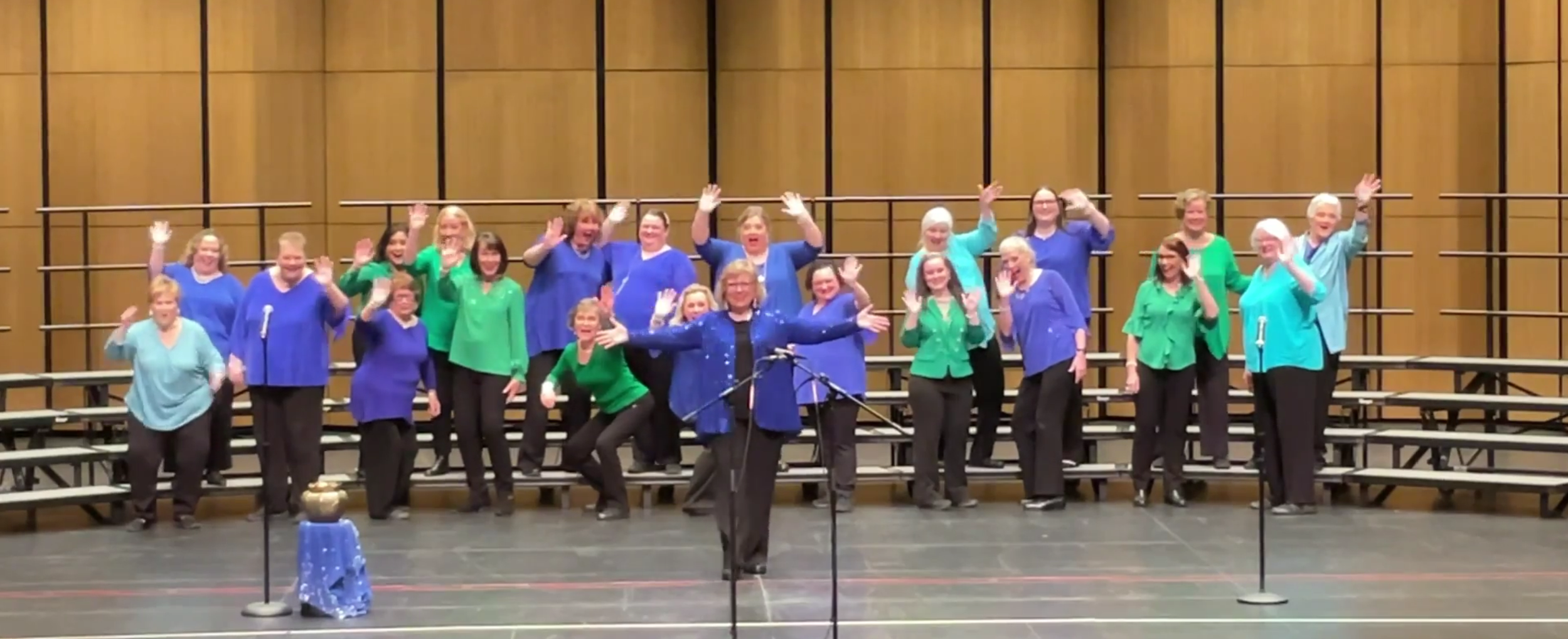 A group of women on a stage, waving and smiling, dressed in multicolored blouses, with some playing percussion instruments.