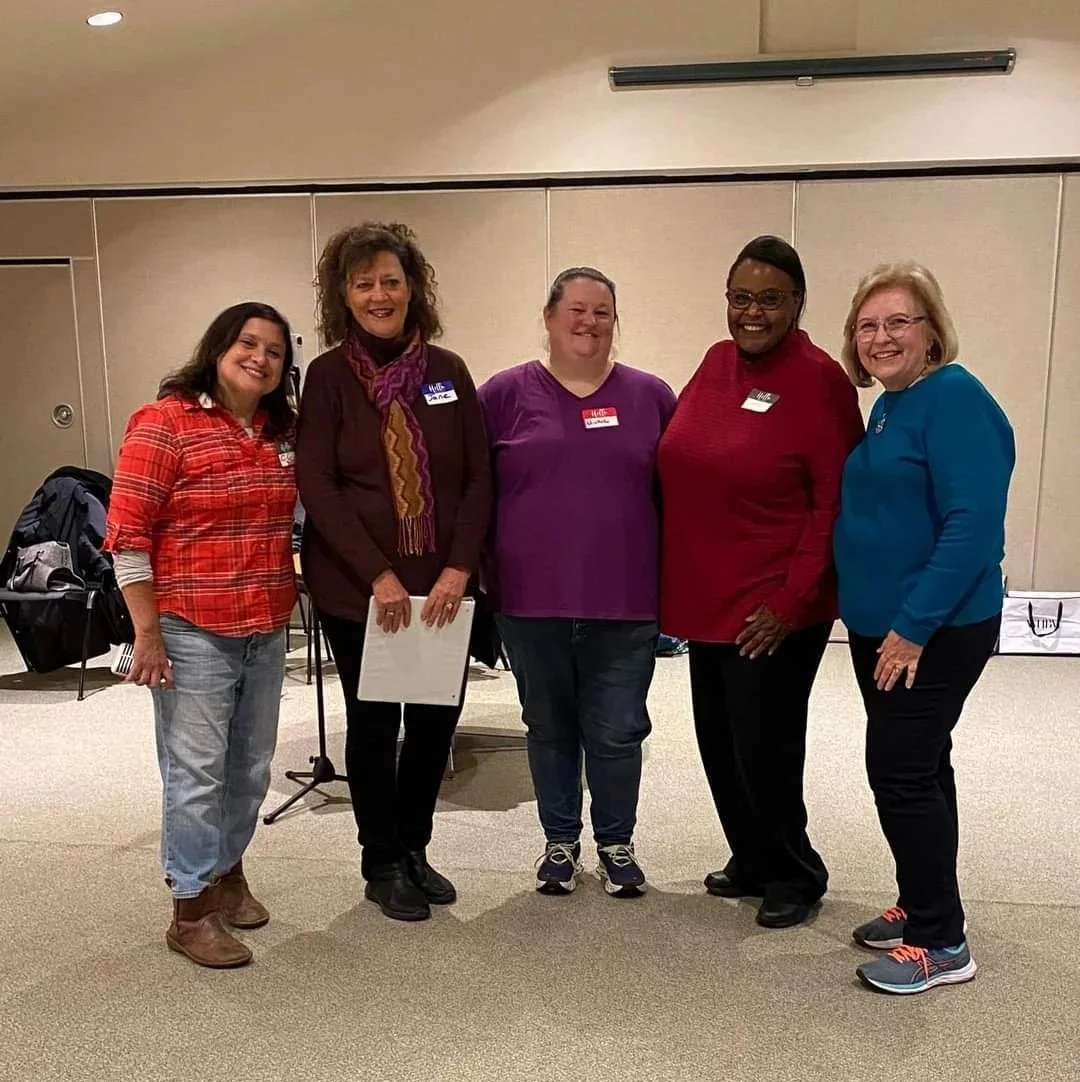 Five women standing together in a room, smiling for a group photo. They are dressed casually and have name tags, possibly at a conference or workshop.