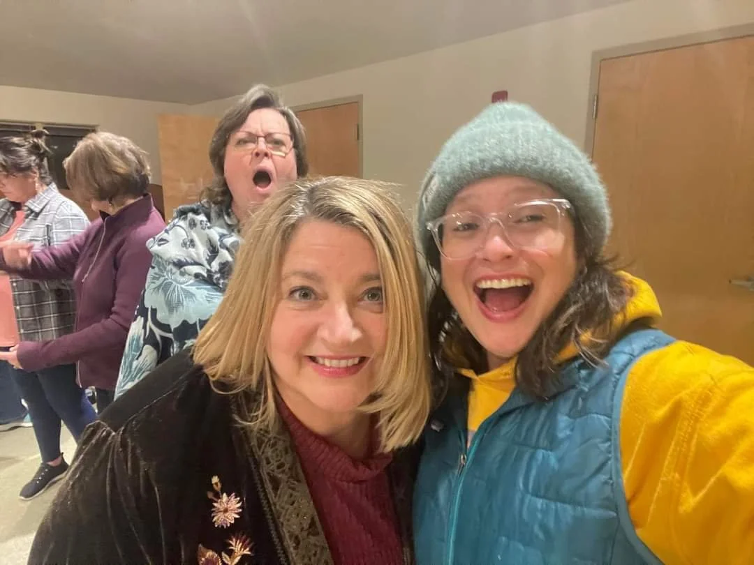 Four women at a social gathering, two in the foreground smiling for a selfie, one in a gray beanie and glasses, yellow jacket, and the other with blonde hair and a dark jacket. Two women in the background appear to be dancing or interacting.