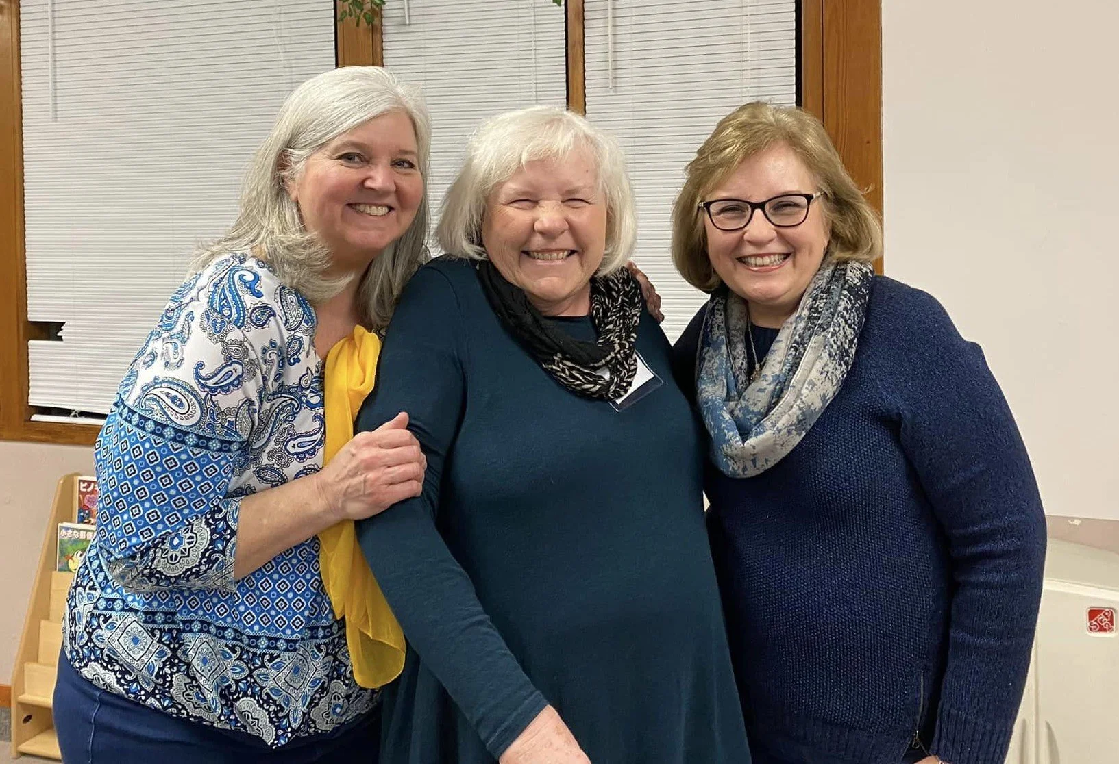 Three women standing together in a room, smiling and hugging.