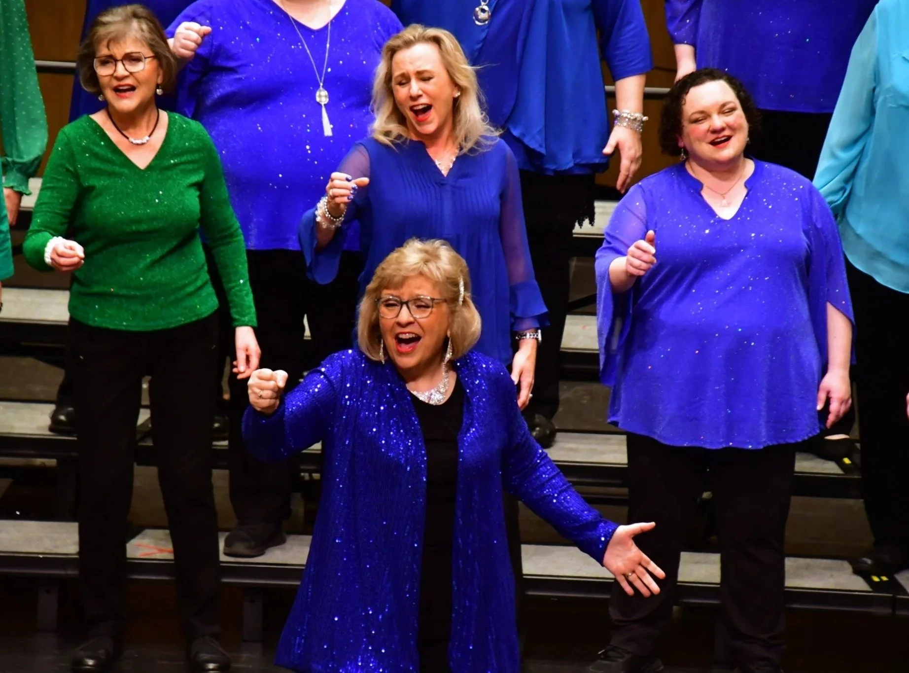 Group of women singing on stage, wearing colorful, sparkly tops, with enthusiastic expressions and gestures.