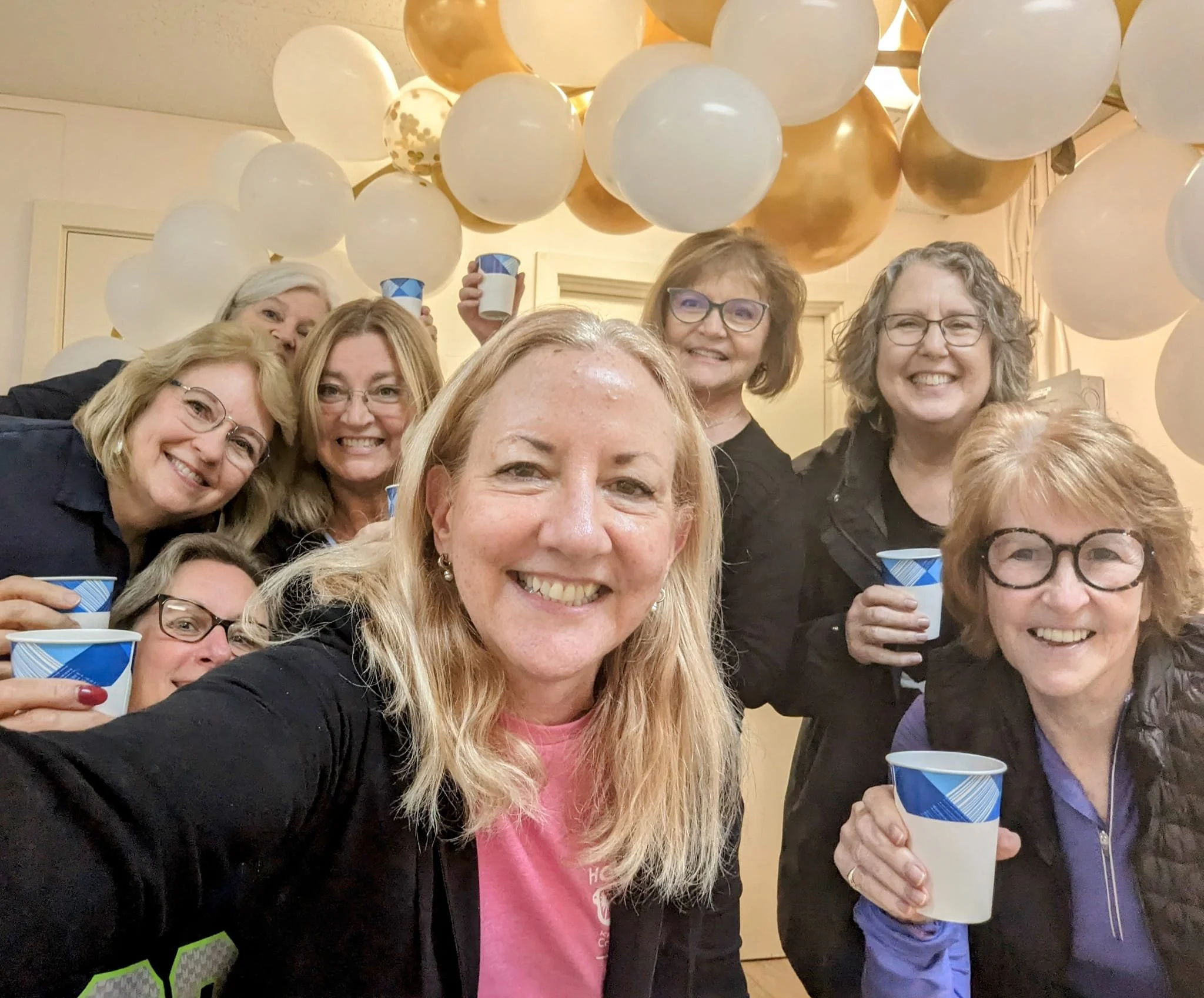 Group of women smiling and celebrating at a party with white and gold balloons, holding cups.