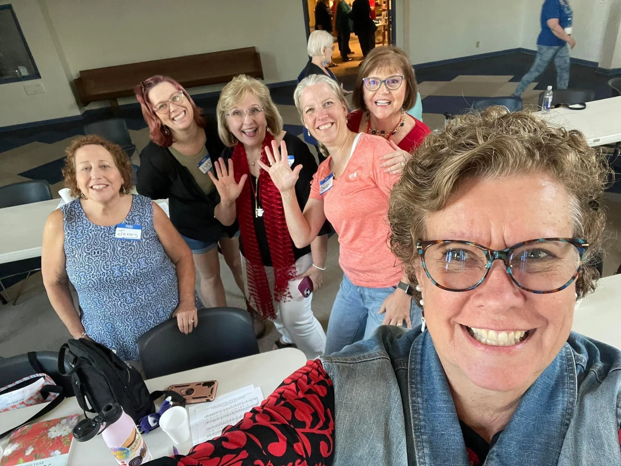Group of women smiling and posing for a selfie at an event, with some wearing name tags, in a room with tables and chairs.