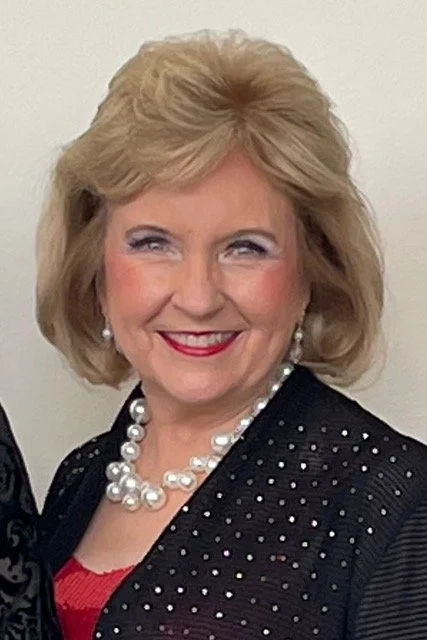 A smiling middle-aged woman with blonde hair wearing pearl earrings, a pearl necklace, and a black jacket with small white dots.