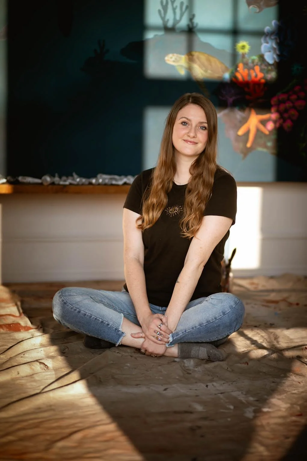 A woman with long brown hair, wearing a black t-shirt and jeans, sitting cross-legged on a wooden floor in front of a blackboard with colorful beach-themed decorations.