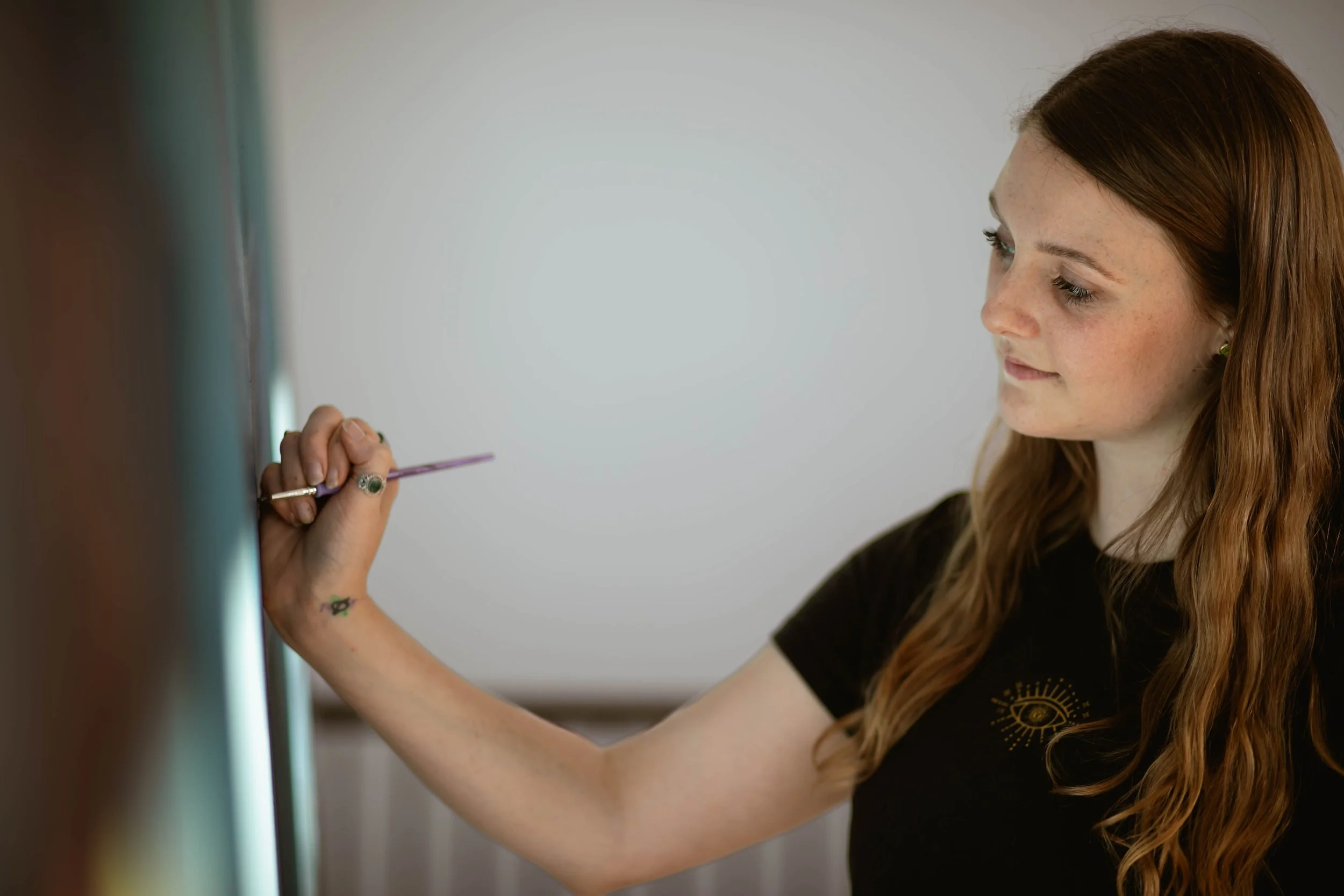 A woman with long wavy red hair and freckles painting on a white surface with a small brush.