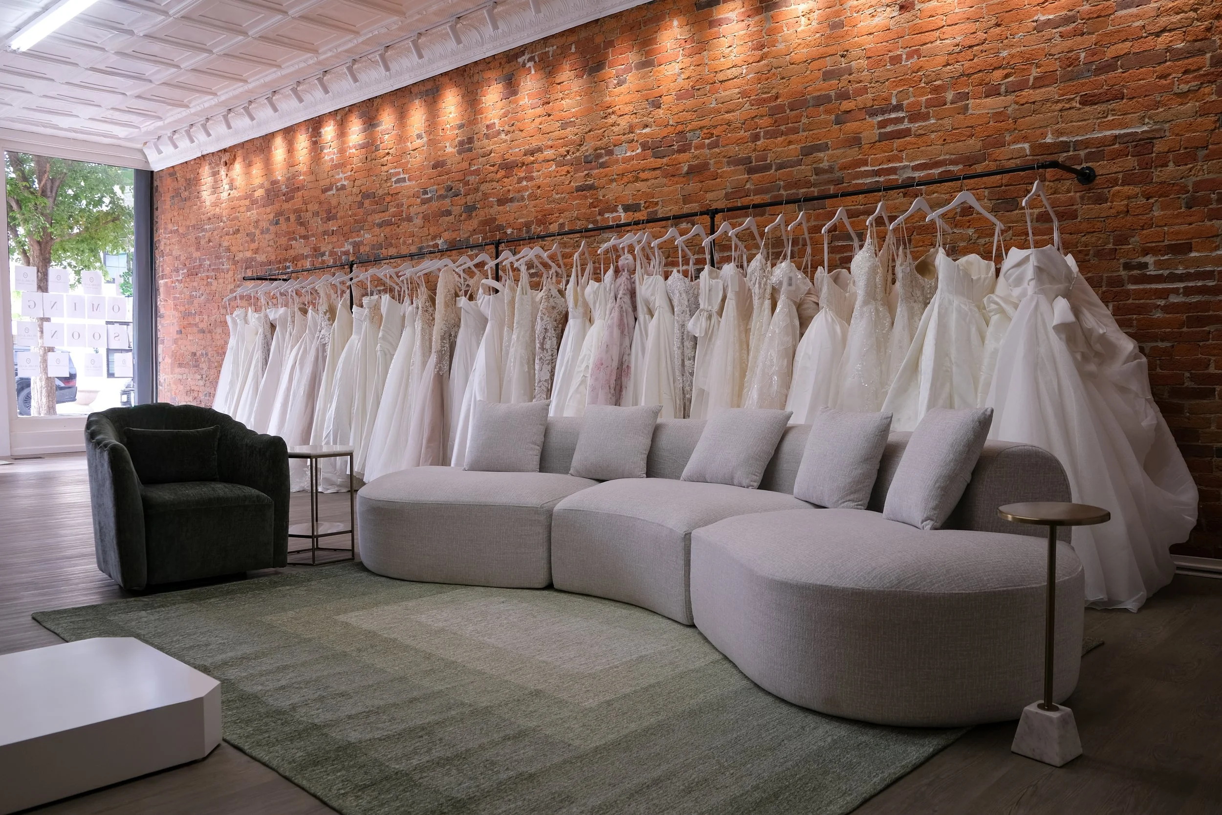 A clothing store displays wedding dresses hanging on a black rack against a brick wall, with a chandelier, gray couch, and chair in the foreground, and a large window showing trees outside.