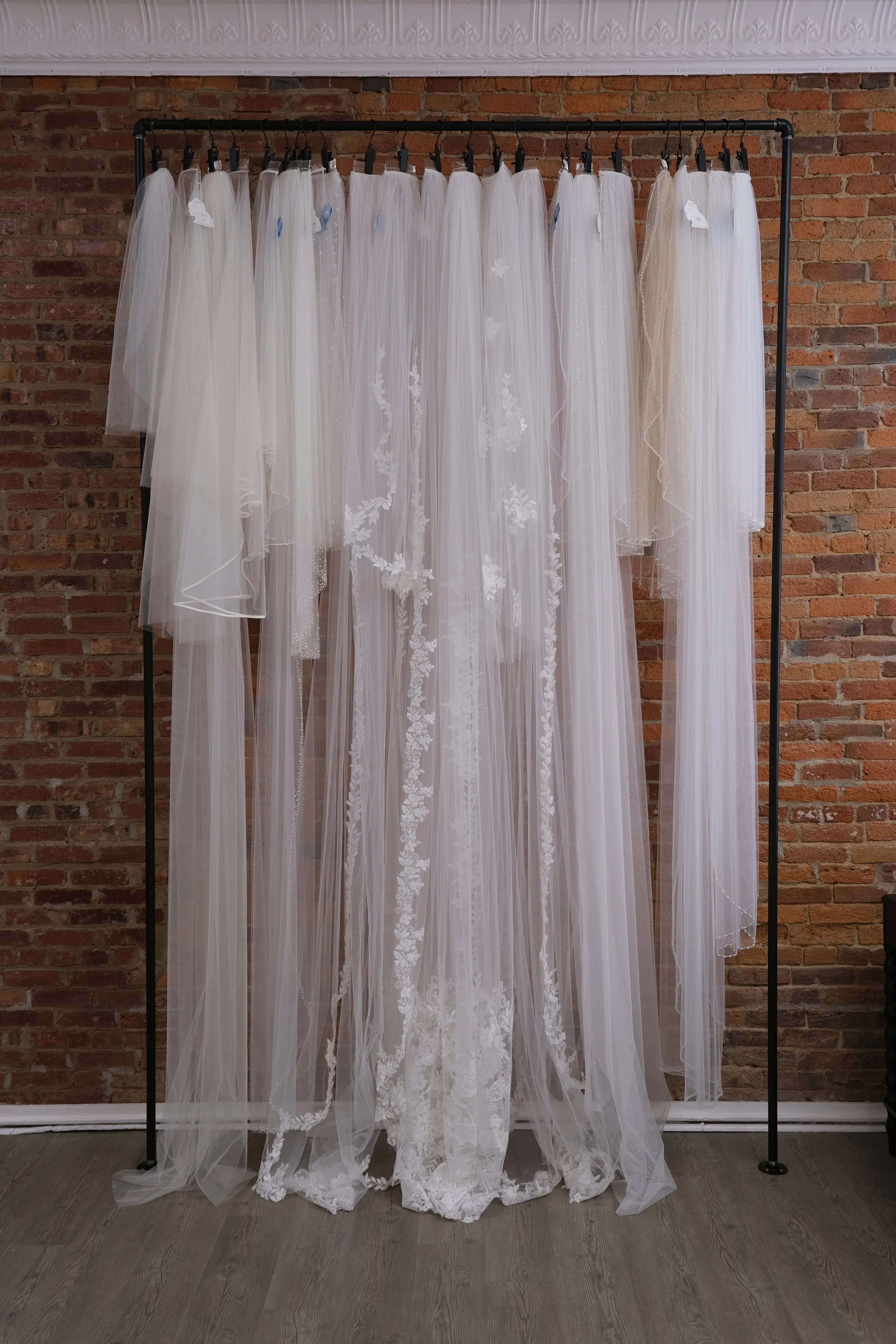 Collection of white bridal wedding dresses hanging on a black clothing rack in front of a brick wall.