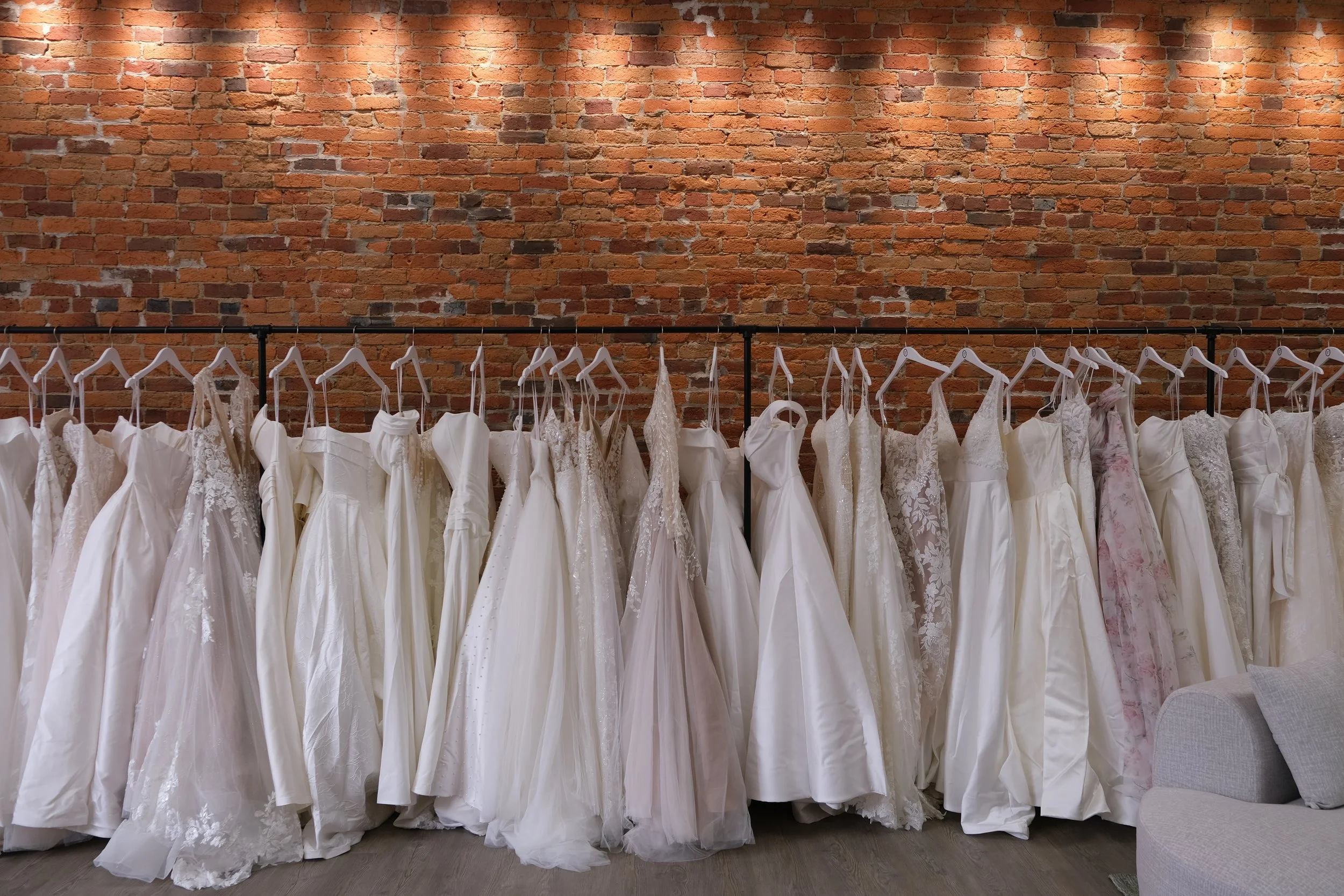 A row of wedding dresses hanging on a black clothing rack in front of an exposed brick wall.