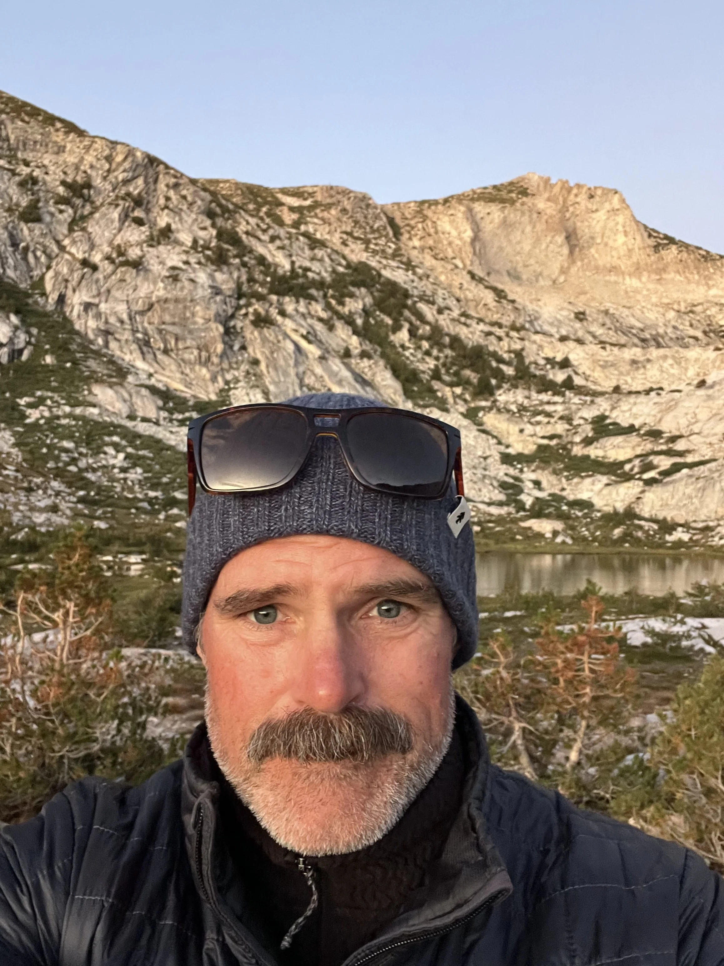 Headshot of Brad Tunis with hat and sunglasses on head standing in front of mountain range
