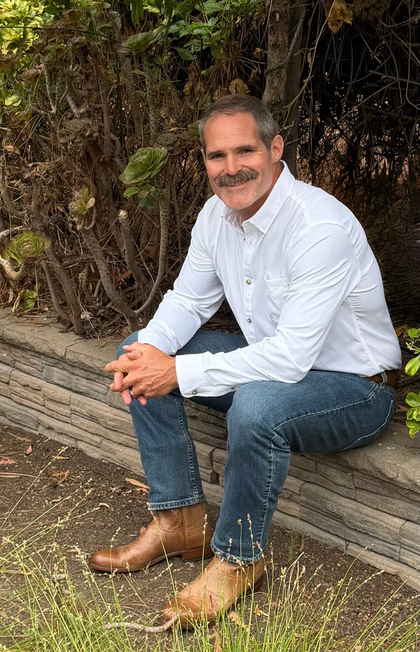 Brad Tunis, hypnotherapist and transformational life coach, wearing a white button-up shirt, jeans, and tan boots, is sitting on a stone bench outdoors amidst plants and foliage, smiling at the camera.