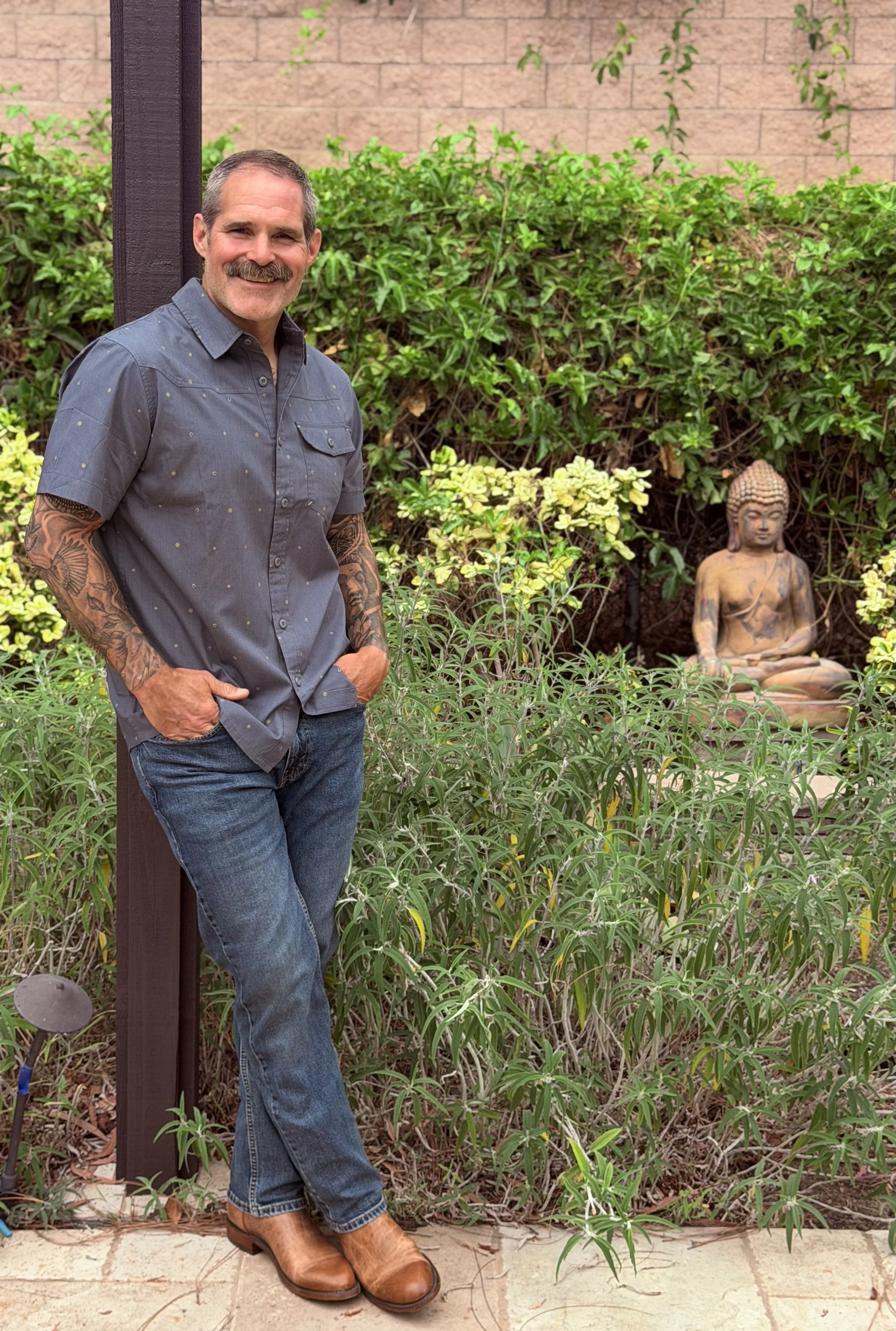 Brad Tunis, hypnotherapist, wearing a blue button-up shirt, jeans, and brown boots, standing outdoors near a garden with a small Buddha statue in the background.