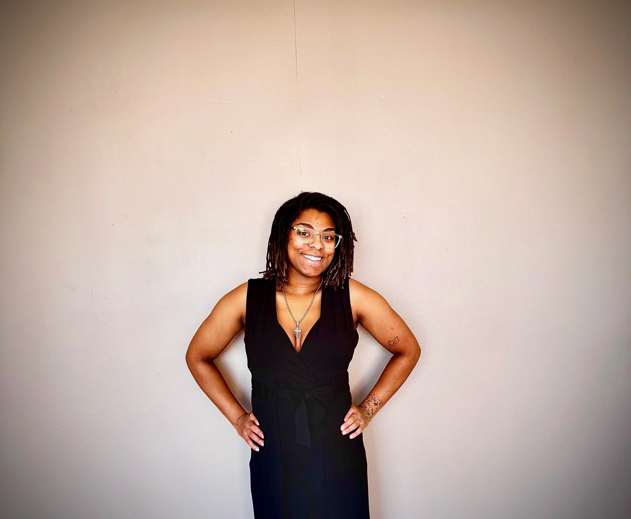 A smiling woman with glasses and dreadlocks is standing against a plain, light-colored wall. She is wearing a black sleeveless top, a necklace with a cross, and has tattoos on her arms.