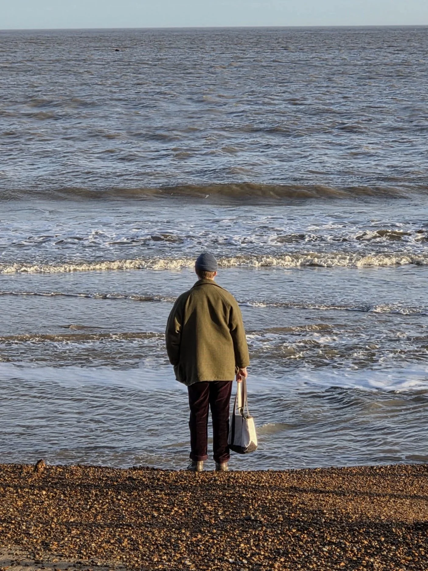 A person standing on a pebbly beach near the ocean, wearing a coat and beanie, holding a tote bag and looking out at the water.