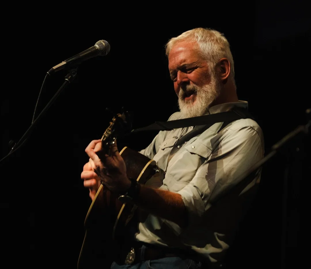 An older man with gray hair and beard playing an acoustic guitar on stage, with a microphone in front of him, against a dark background.