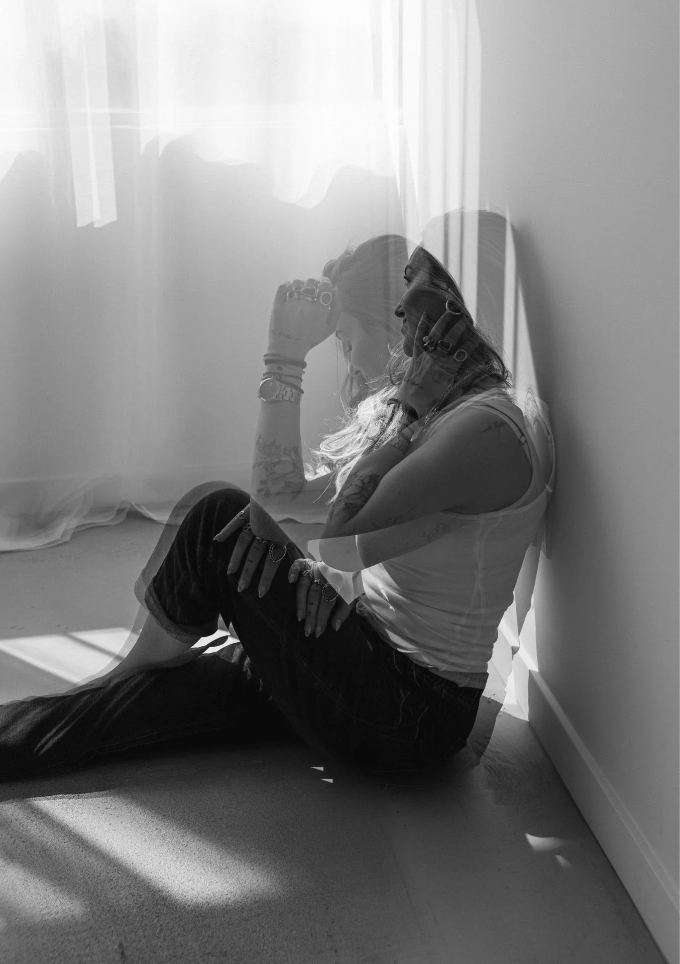 A woman with long hair sitting on the floor against a wall, appearing to be in a reflective or contemplative state, with shadows falling across her and the wall.