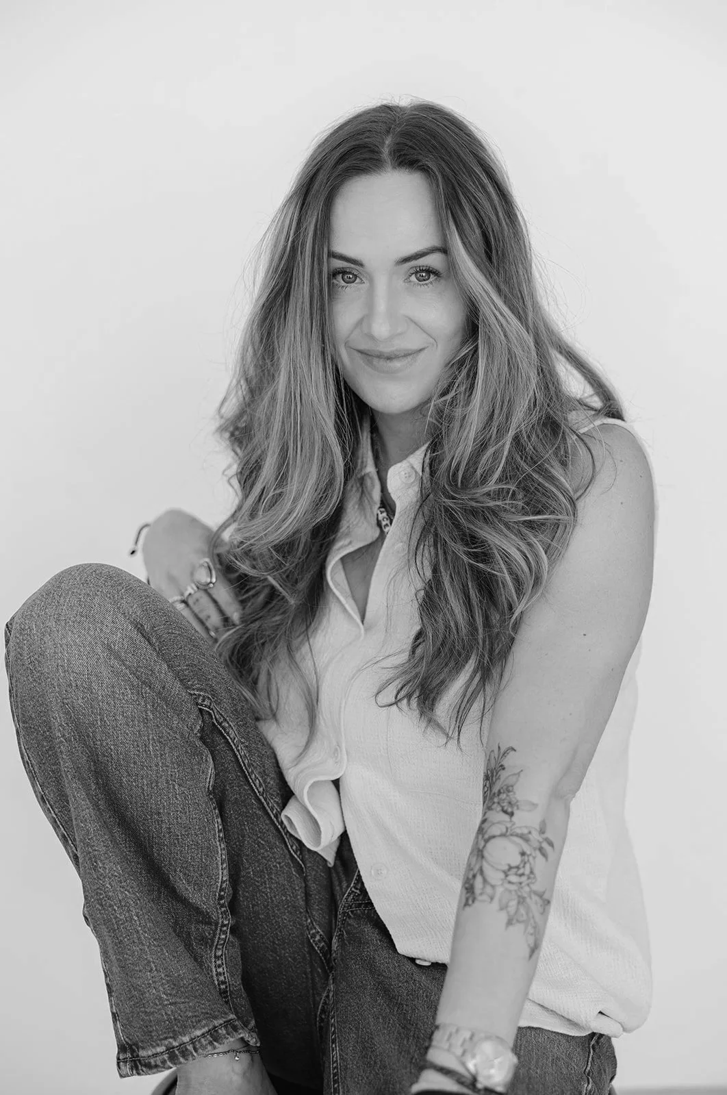 Black and white photo of a woman with long wavy hair, sitting with one leg crossed. She is wearing a sleeveless shirt, denim pants, and accessories including a watch and bracelet. She is smiling slightly and looking at the camera.