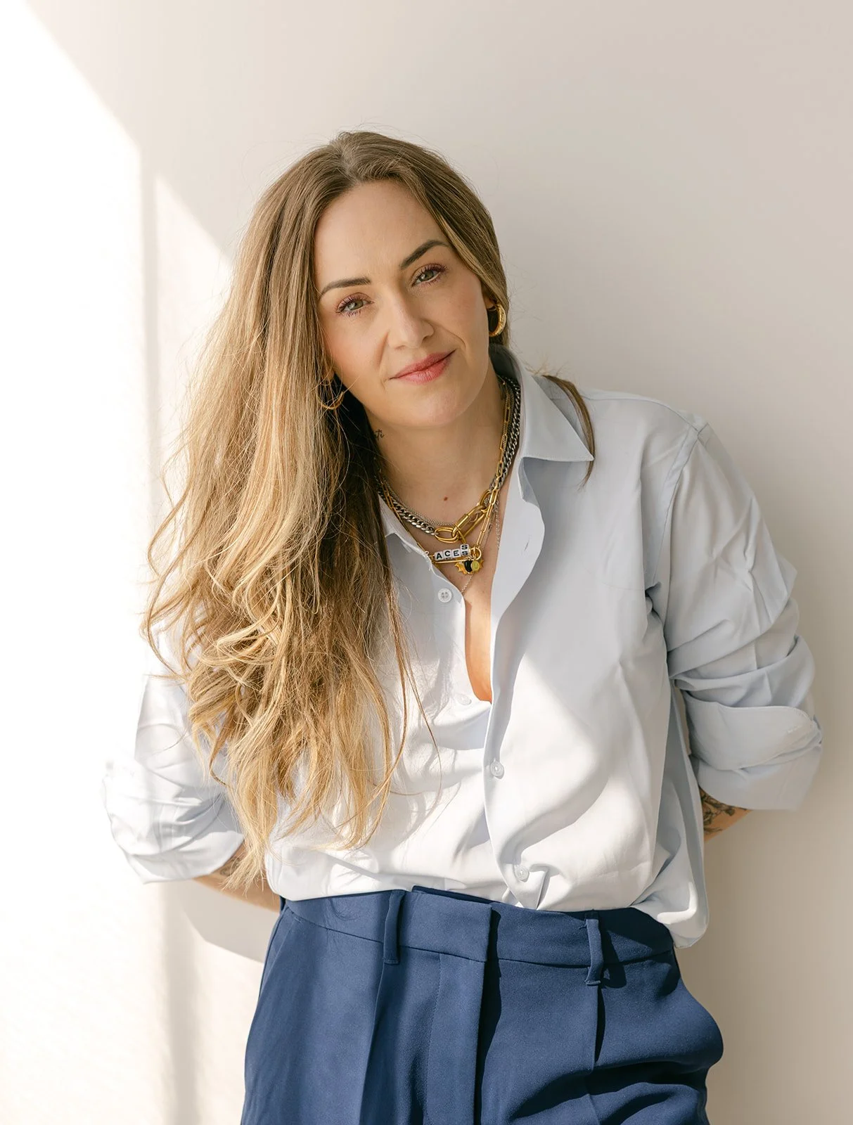 A woman with long, wavy brown hair wearing a white button-up shirt and navy blue tie-waist pants, accessorized with layered gold jewelry, standing against a light-colored wall.
